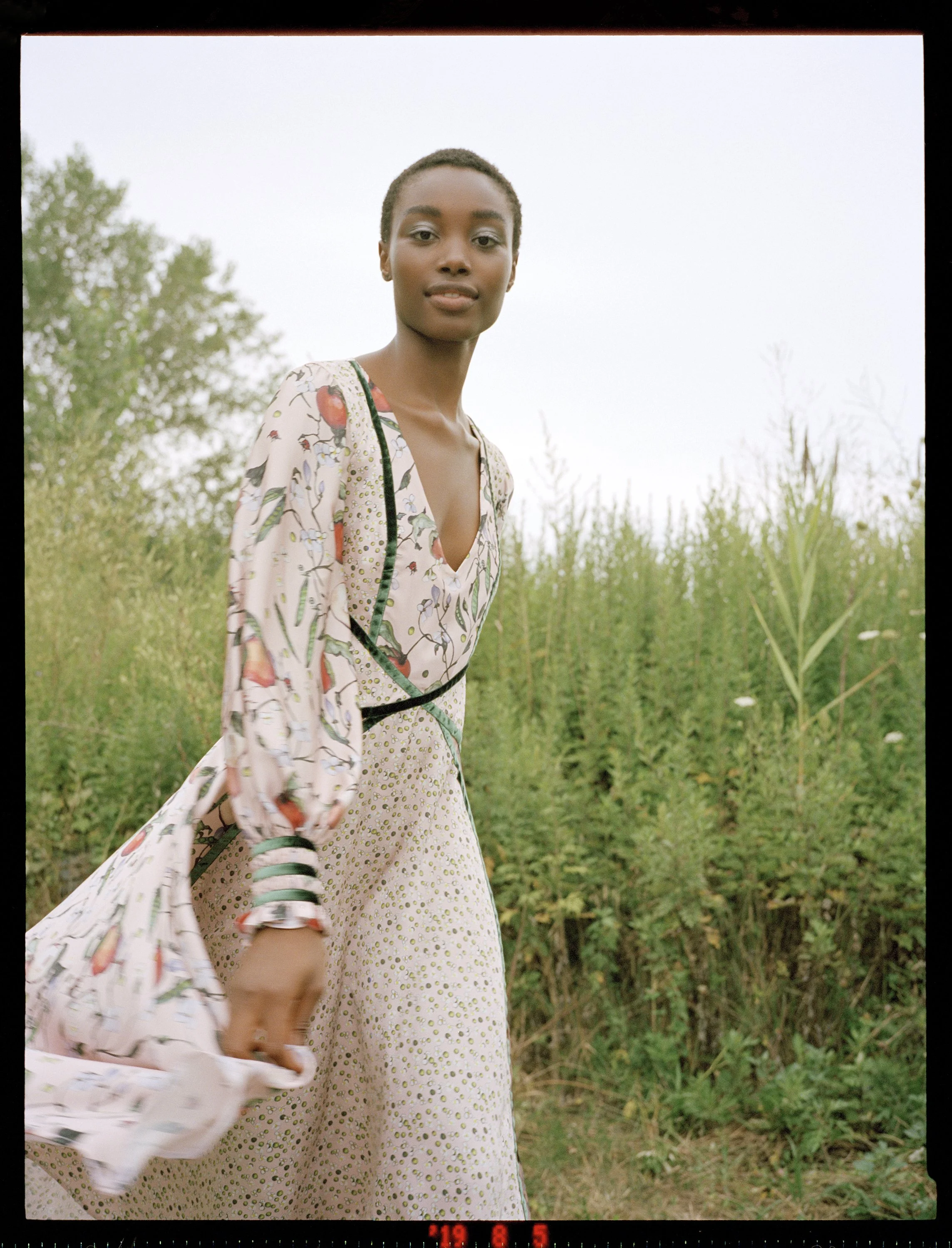 A young woman in a patterned dress standing in a green field during daytime.