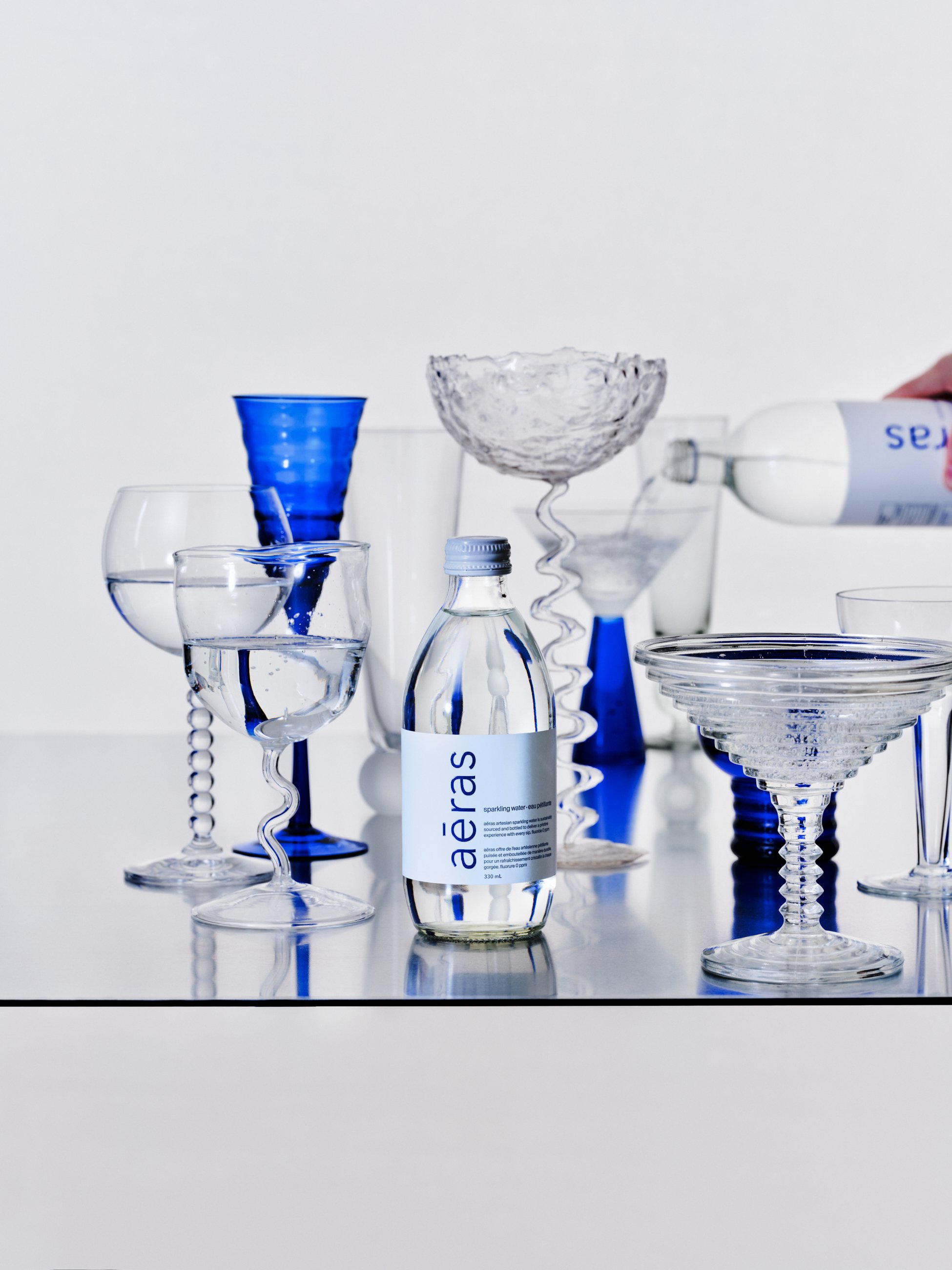 Various glassware and a bottle of Aeás sparkling water on a mirror surface, with some bottles pouring water.