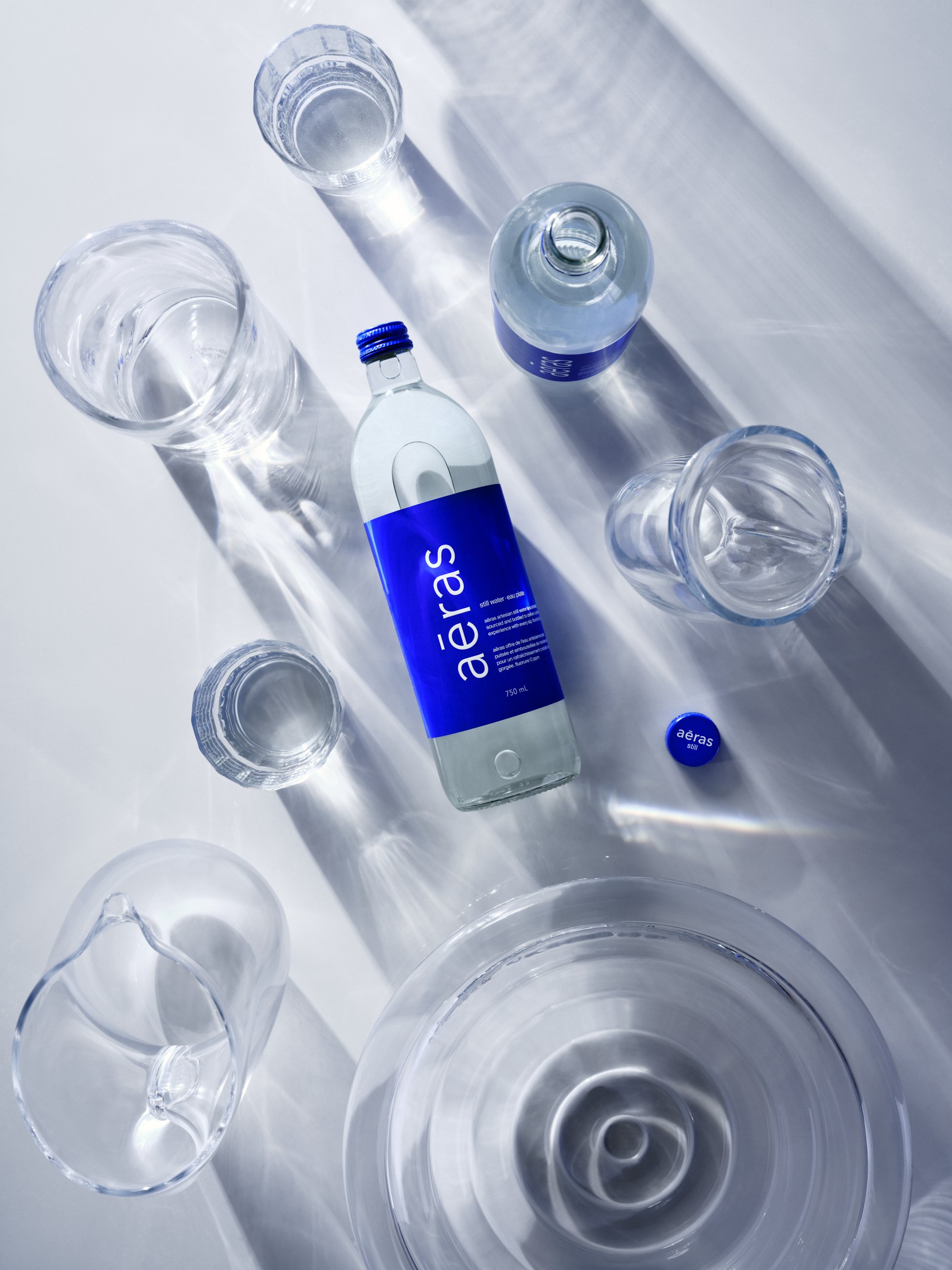 A bottle of Aeras seawater with four empty glasses, a small water bottle, a bowl, and a round blue object on a white surface with sunlight creating shadows.