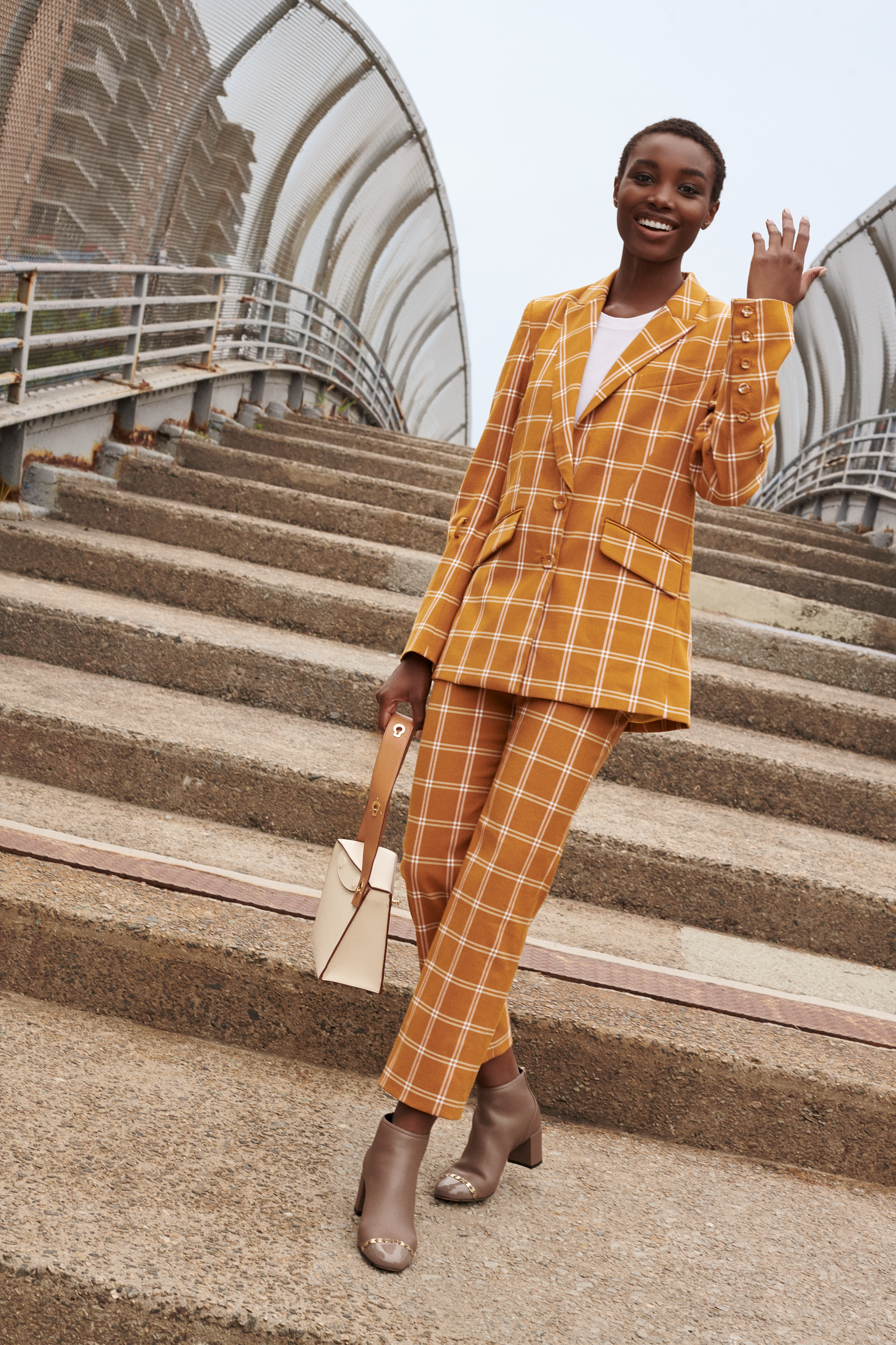 A woman in a yellow checkered suit and taupe ankle boots smiling and waving on outdoor stairs.