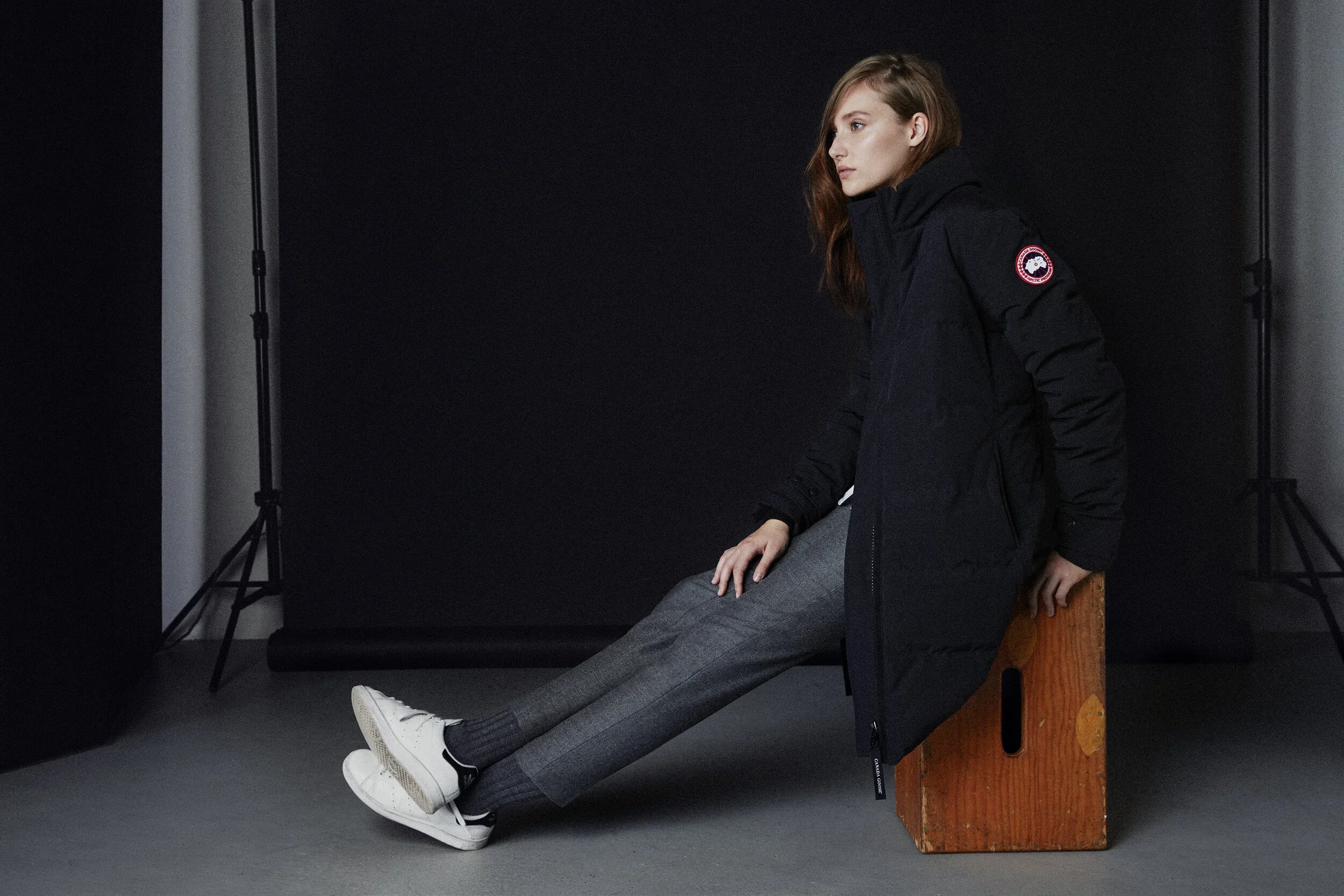 A woman with red hair sitting on a wooden box against a black background, wearing a black jacket, gray pants, and white sneakers.