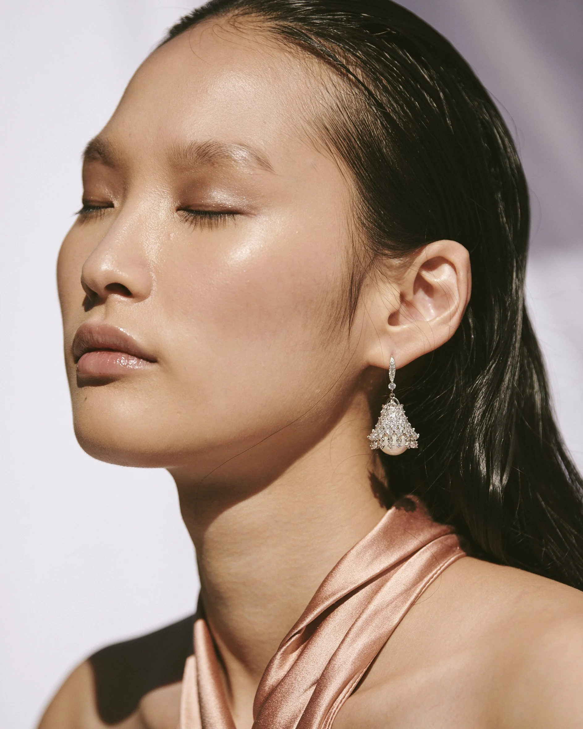 Close-up of a woman with closed eyes, light makeup, and wet black hair wearing a pink satin top and a dangling diamond earring.