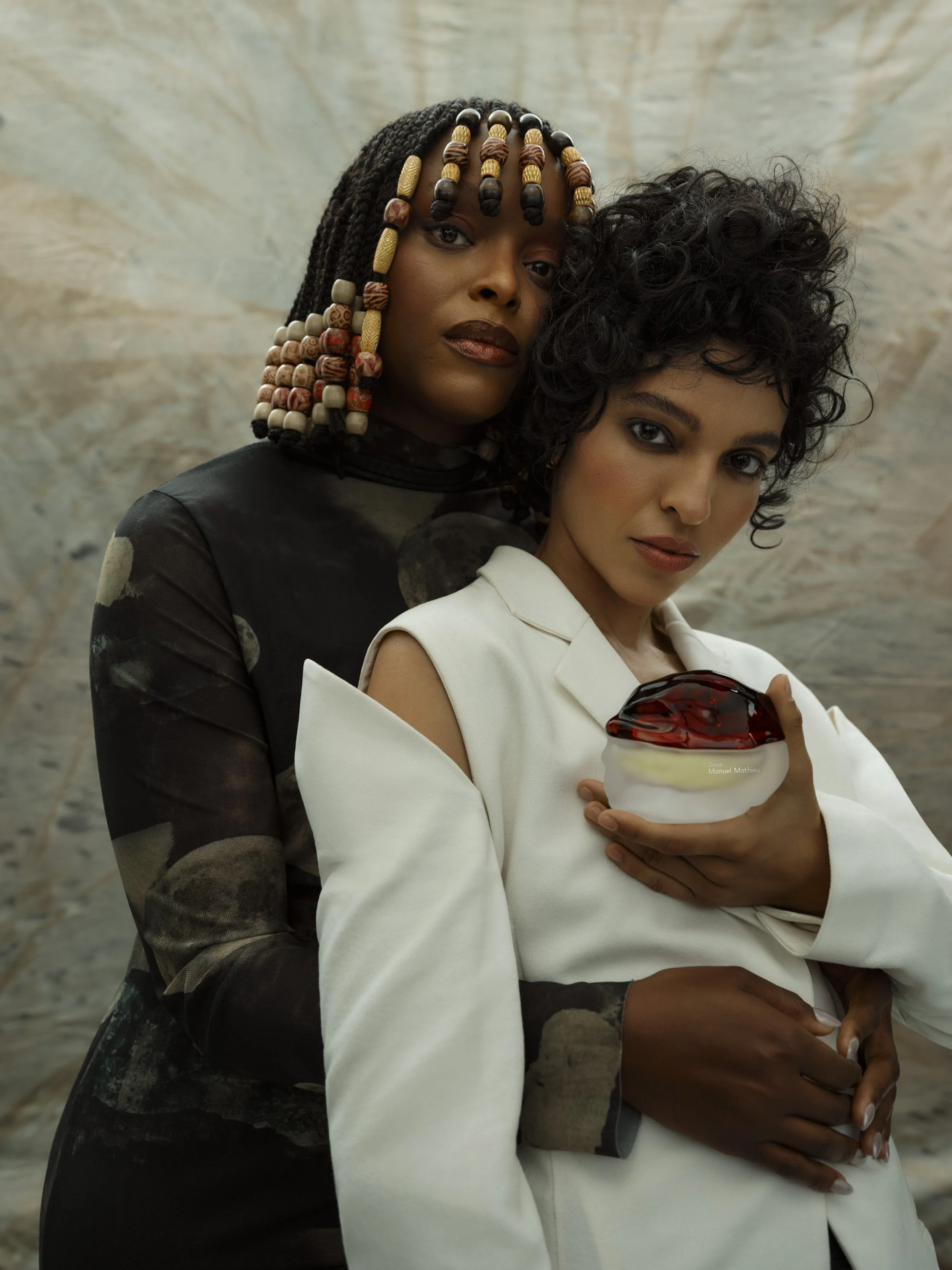Two women pose together, one with beaded braided hair and the other with short curly hair, holding a glass bowl with a layered dessert. They stand against a modern, abstract background.