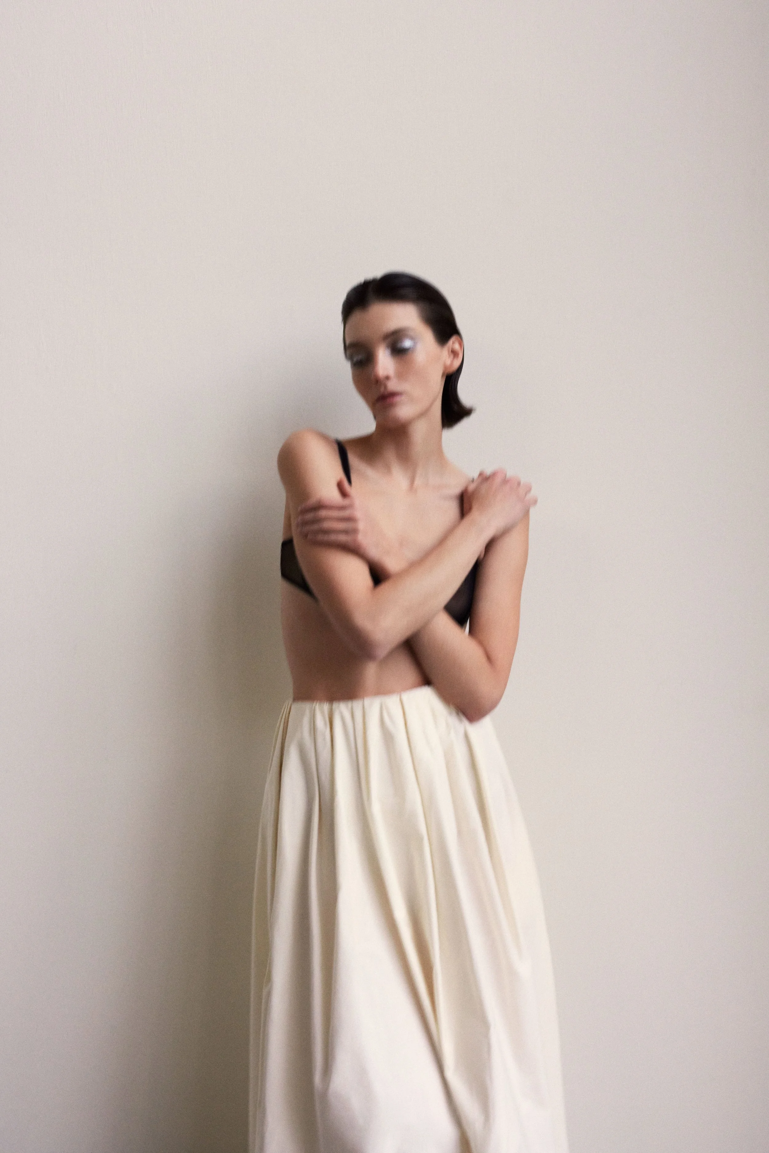 Woman with dark hair and makeup, wearing a black bra and cream-colored skirt, standing against a plain white wall with arms crossed.