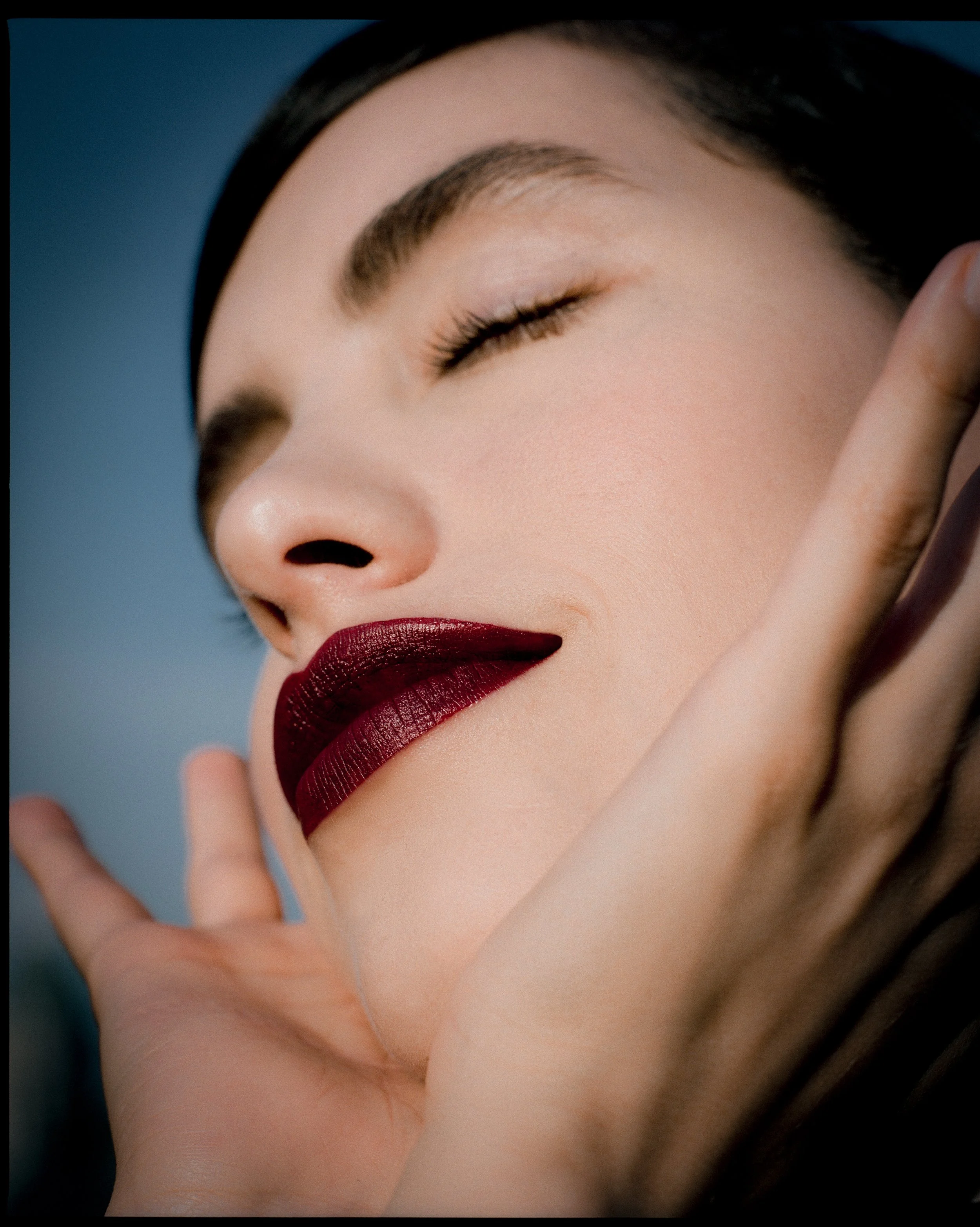 Close-up of a woman's face with closed eyes, dark hair, bold dark red lipstick, and a peaceful smile, gently touching her face.