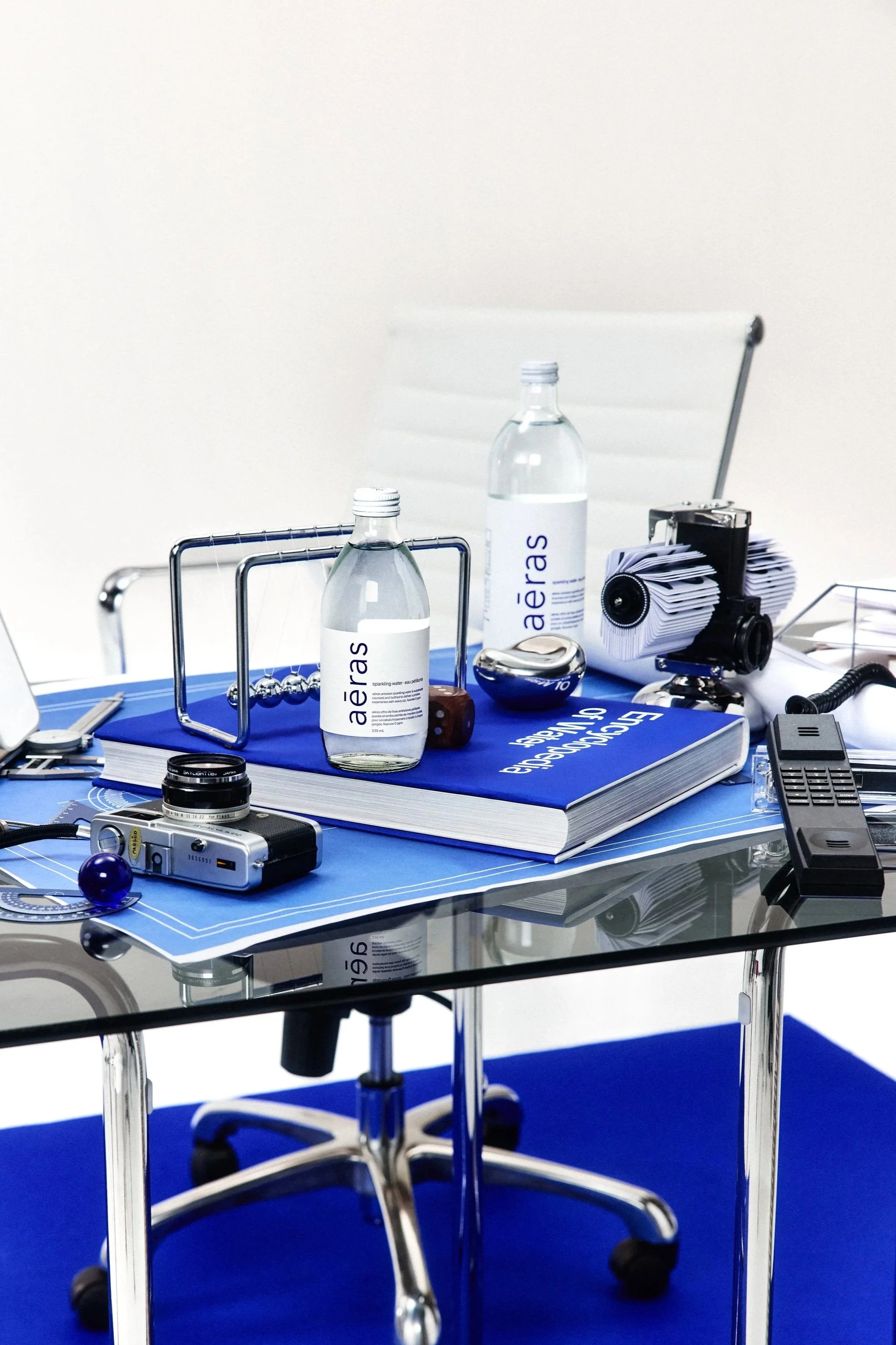 A cluttered desk with various items including two bottles of water labeled 'aerA's', a notebook, a microscope, a vintage camera, a remote control, a handmade Newton's cradle, and other miscellaneous objects, on a glass table with a blue rug underneat