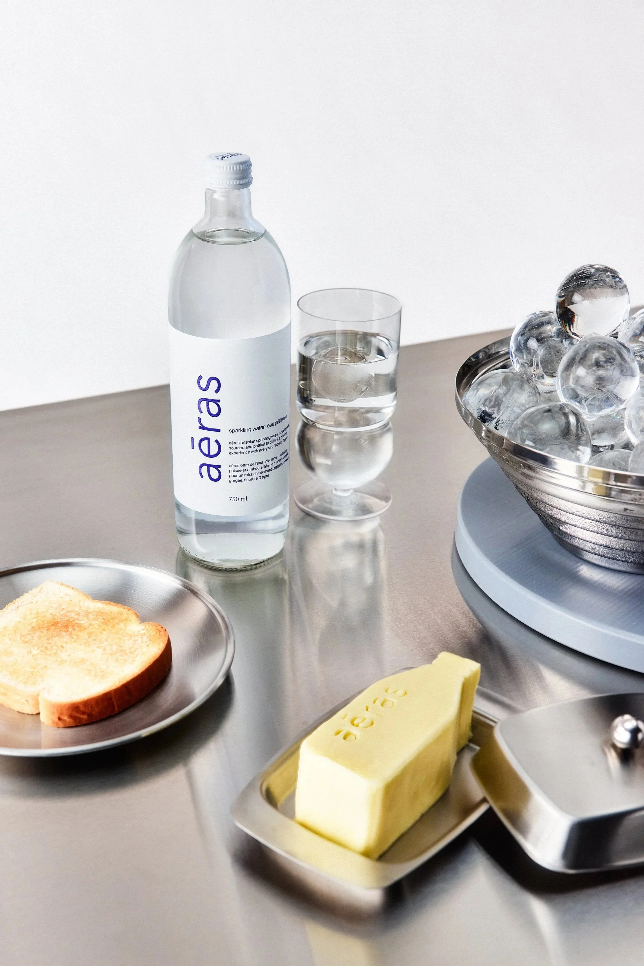 Bottle of sparkling water, glass of water, bowl filled with ice cubes, butter, toast on a plate, on a stainless steel table.