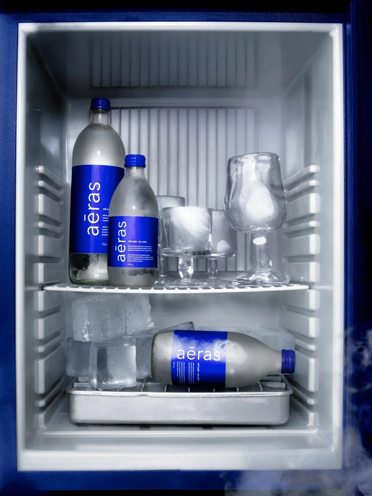 The image shows a refrigerator with bottles of Aero water, ice cubes, and glassware on two shelves.