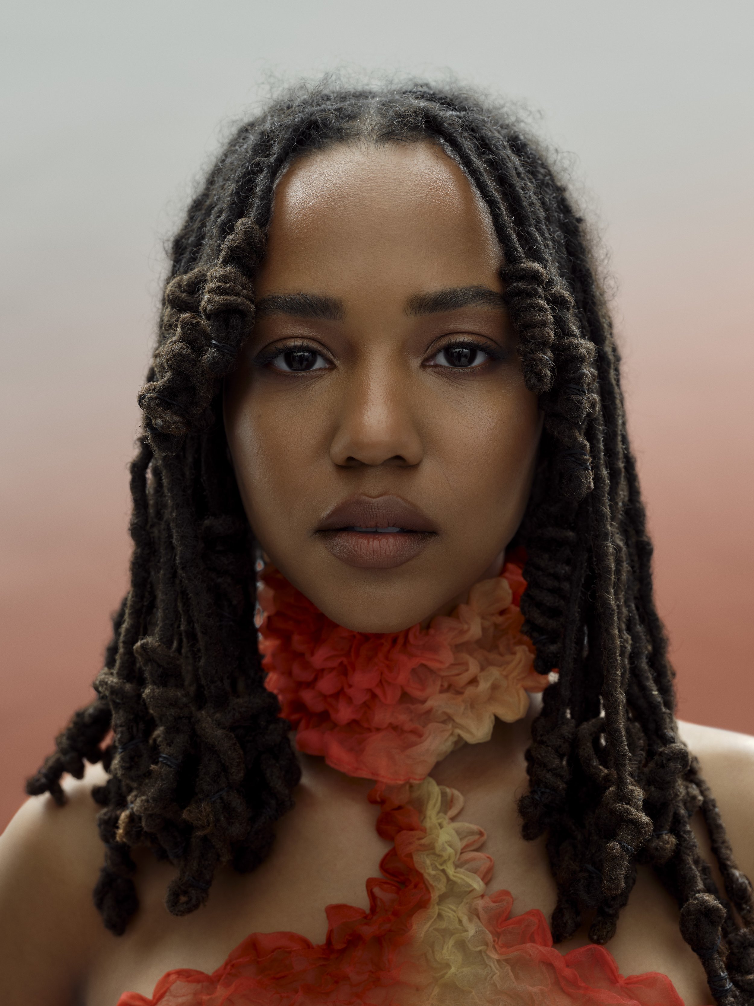 Close-up of a woman with dark, textured dreadlocks and natural makeup, wearing a ruffled, multicolored neckpiece, against a soft gradient background.