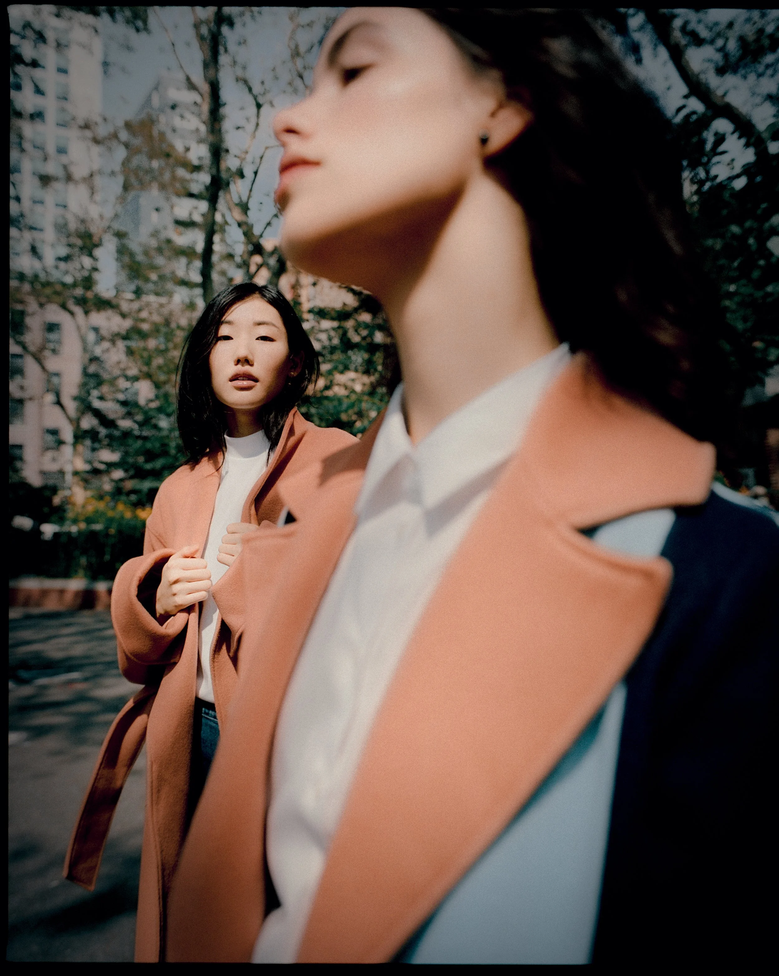Two women outdoors in a city park with trees and buildings in the background. The woman in the foreground has dark hair, a white shirt, and a tan coat, and is only partially visible. The woman in the background has dark hair, and is wearing a tan coa