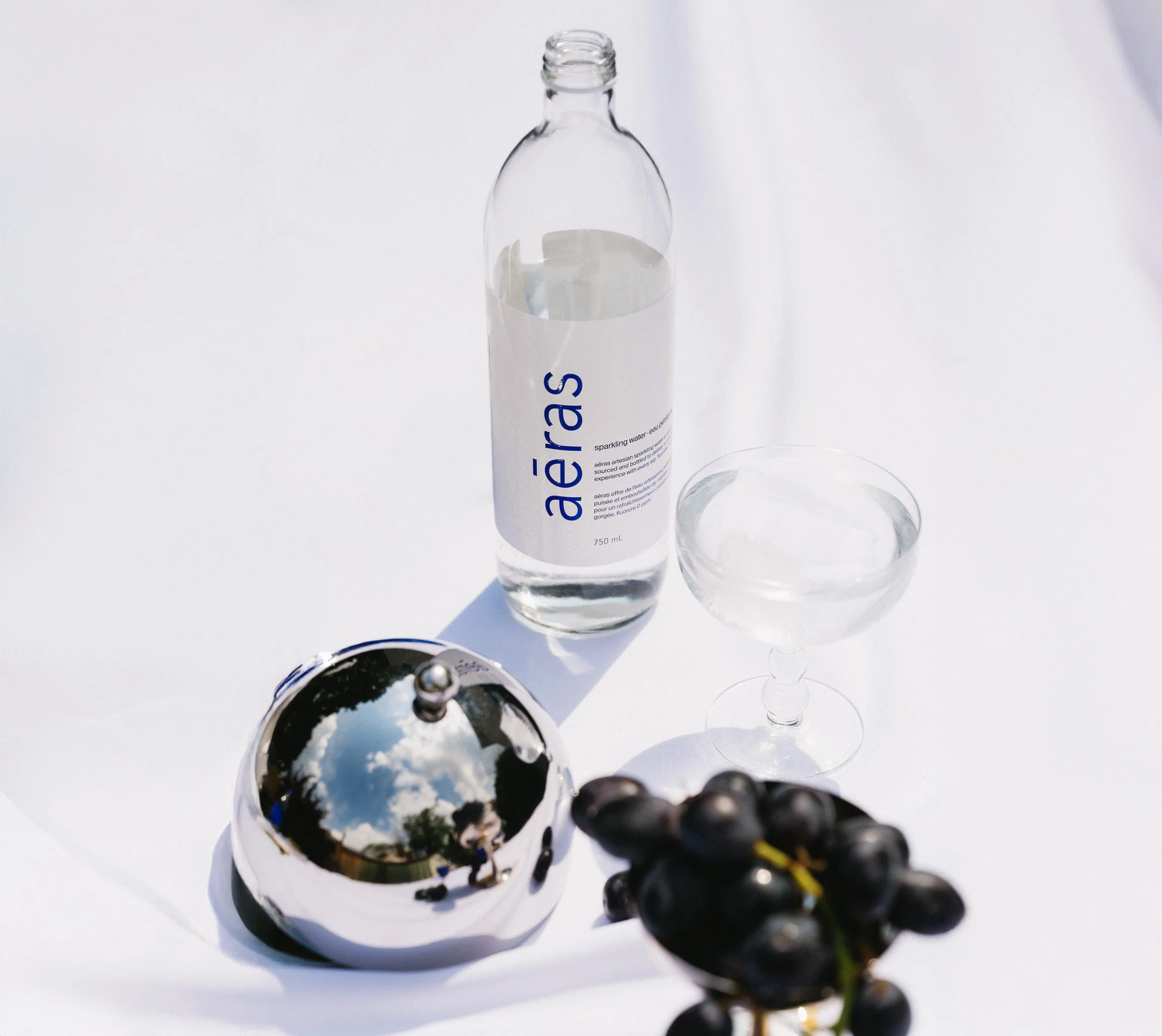 A bottle of sparkling water labeled 'aéras', a coupe glass, a bunch of black grapes, a small reflective spherical object, and their shadows on a white surface.