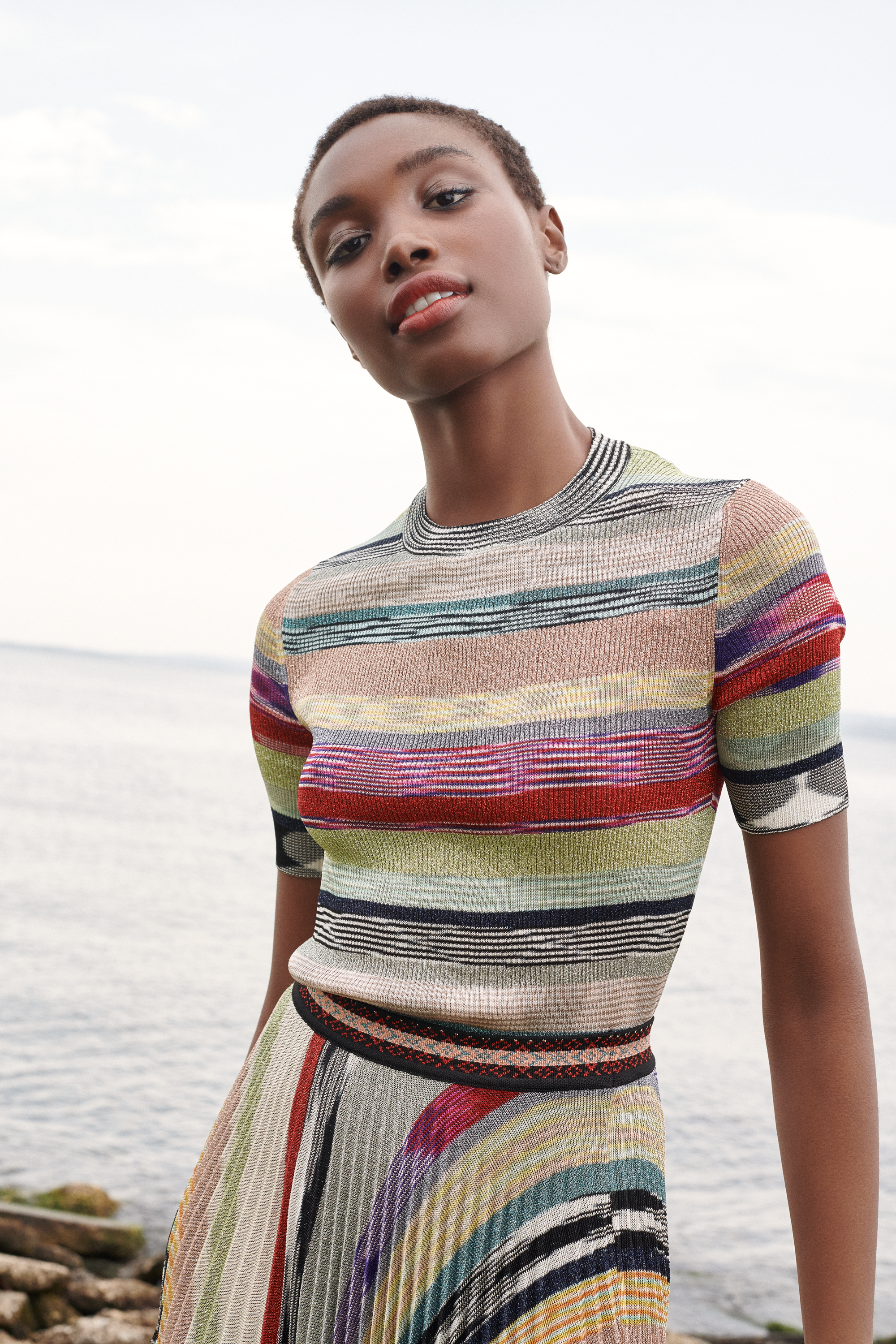A woman outdoors near water, wearing a colorful, striped knit dress, standing with a slight tilt of her head.