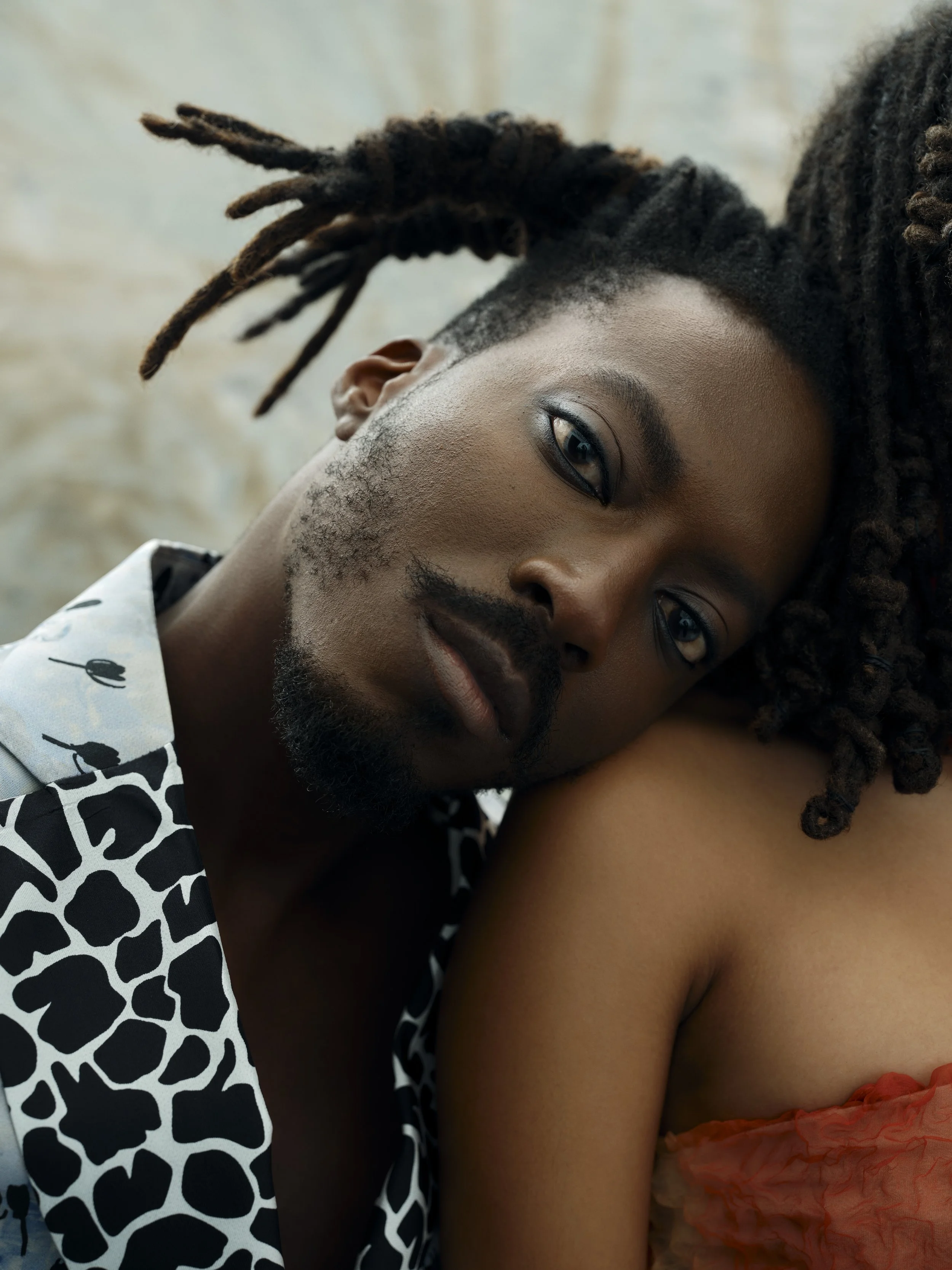 Close-up of a man with dreadlocks resting his head on a woman's shoulder, both looking into the camera.