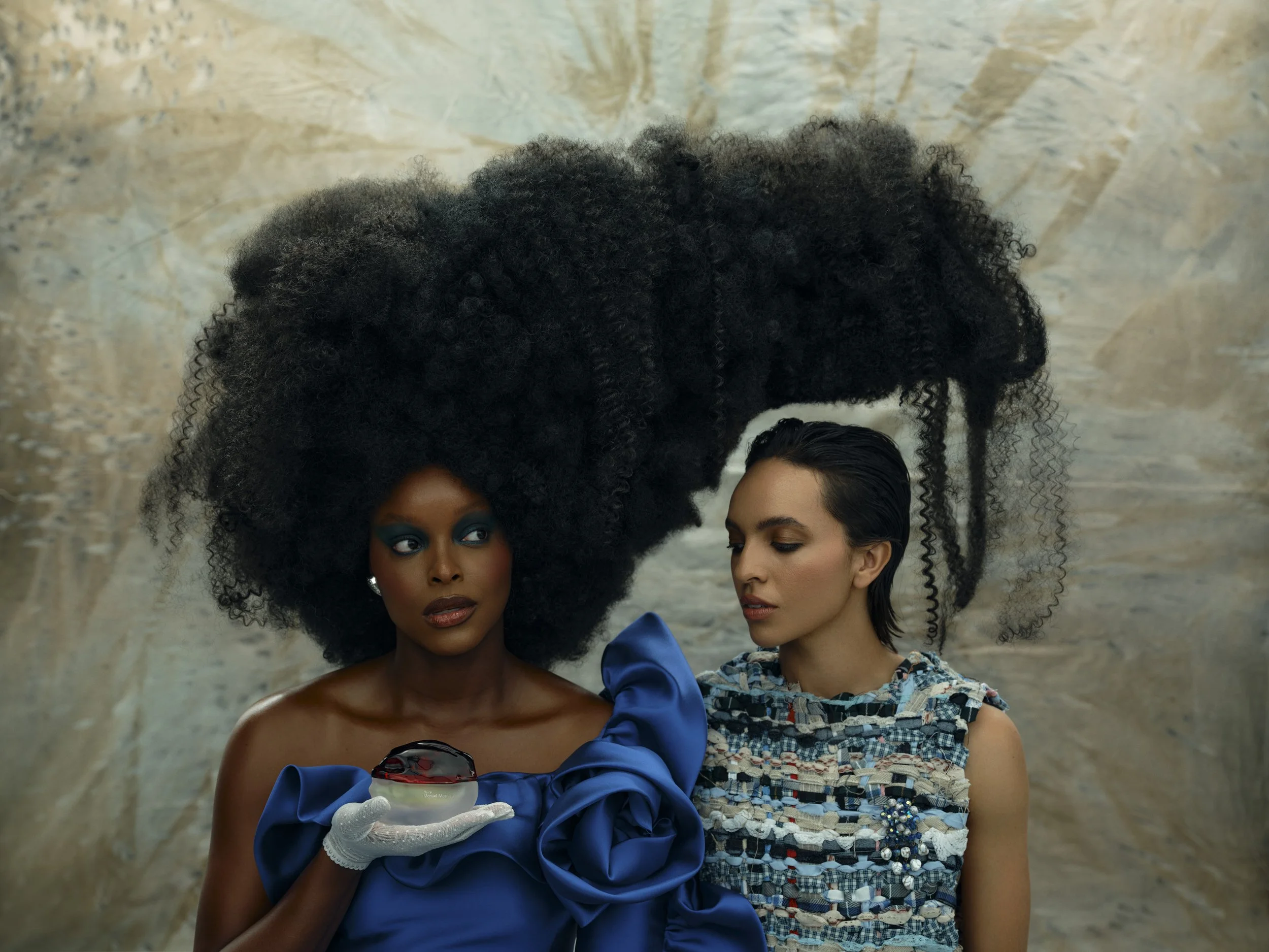 Two women standing together, one with an elaborate large afro hairstyle and wearing a blue dress with puffed sleeves, holding sunglasses, and the other with slicked-back hair wearing a multicolored, textured dress, in front of a neutral marble backgr