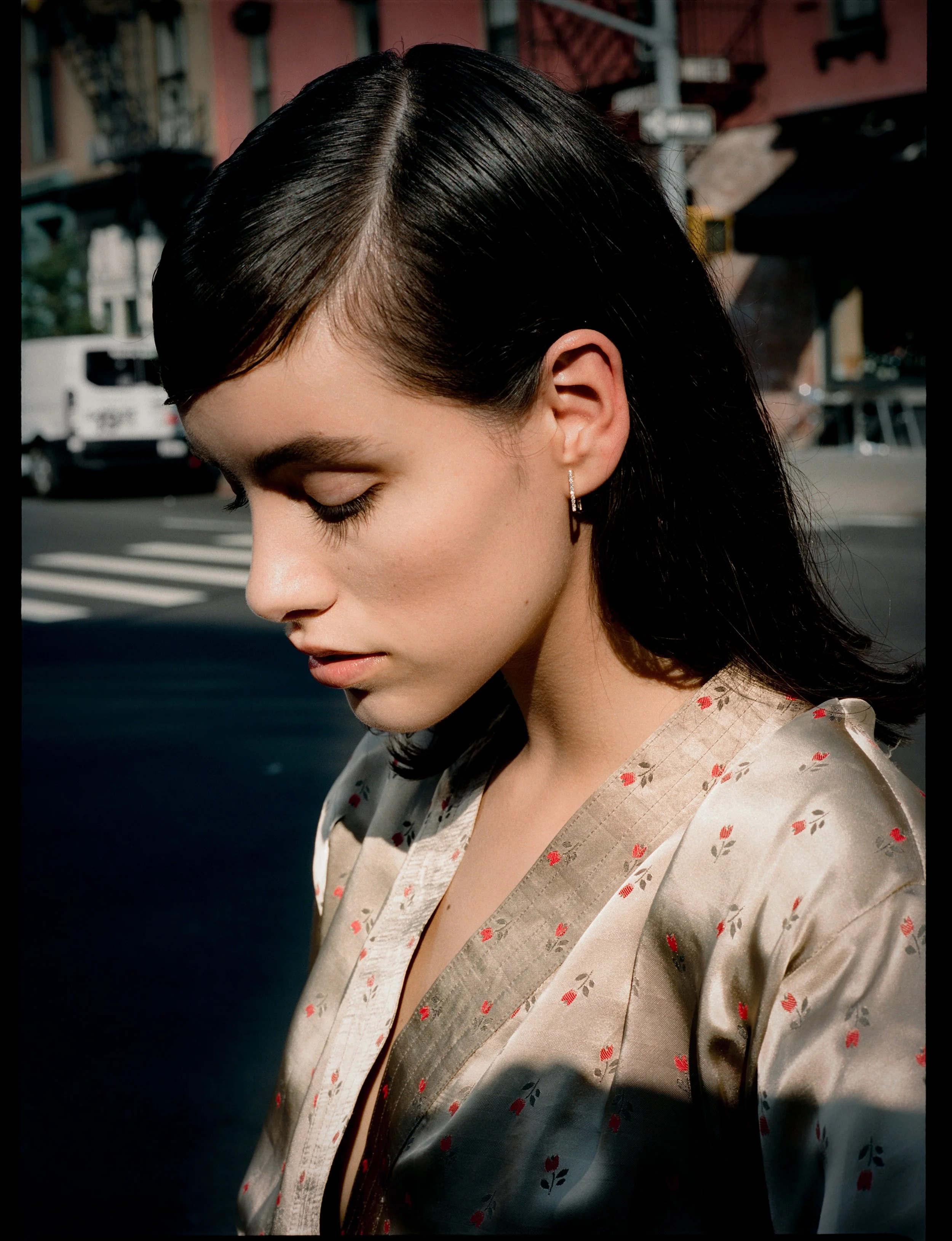 A woman with dark, straight hair and earrings looks down, wearing a light-colored silk robe with small red and green floral patterns. She is outdoors on a city street with buildings, parked cars, and a crosswalk in the background.