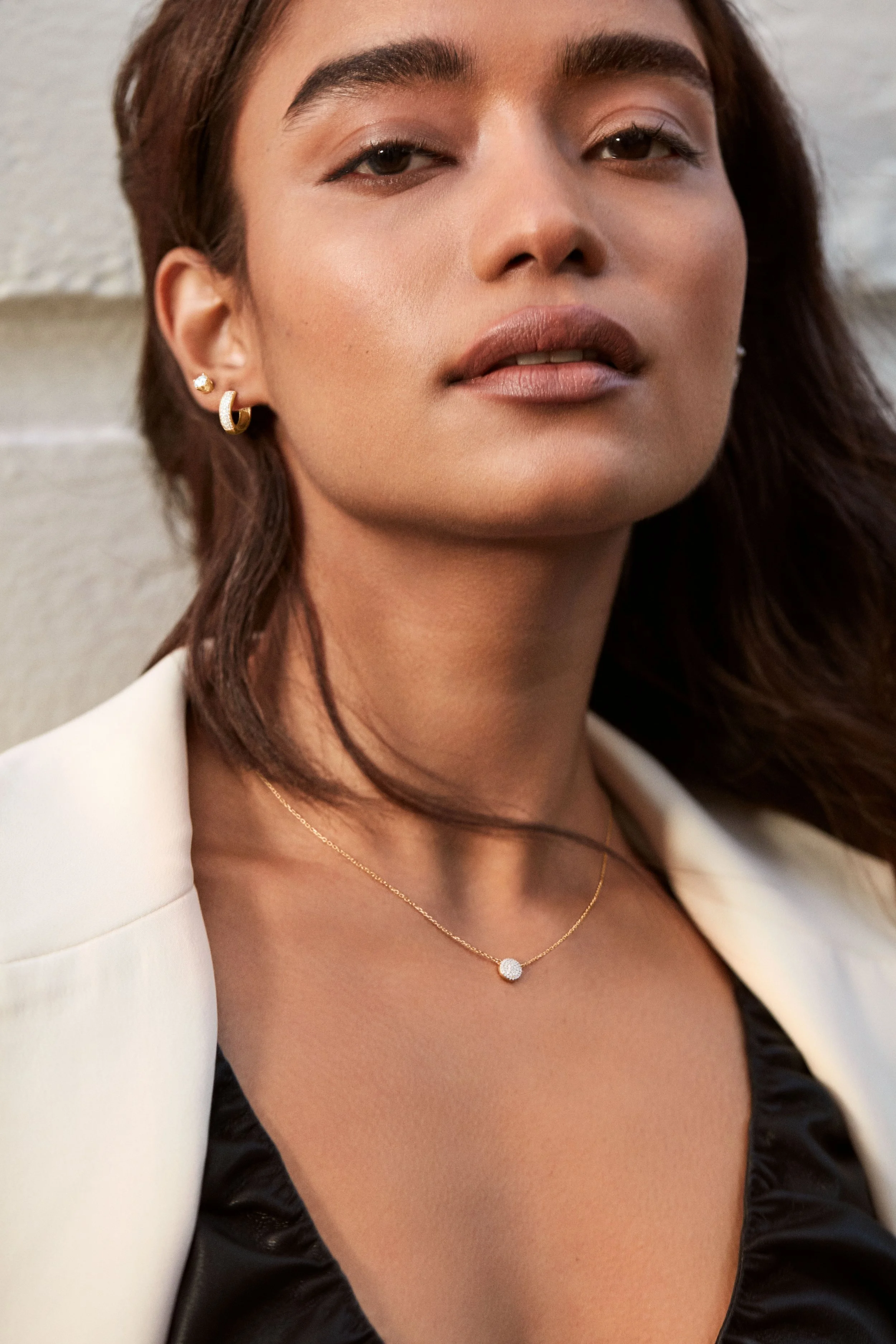 A woman with tan skin and dark brown hair, wearing earrings and a delicate gold necklace with a small round pendant. She is dressed in a black top and a white blazer, with a neutral expression and a slightly tilted head, standing against a light-colo