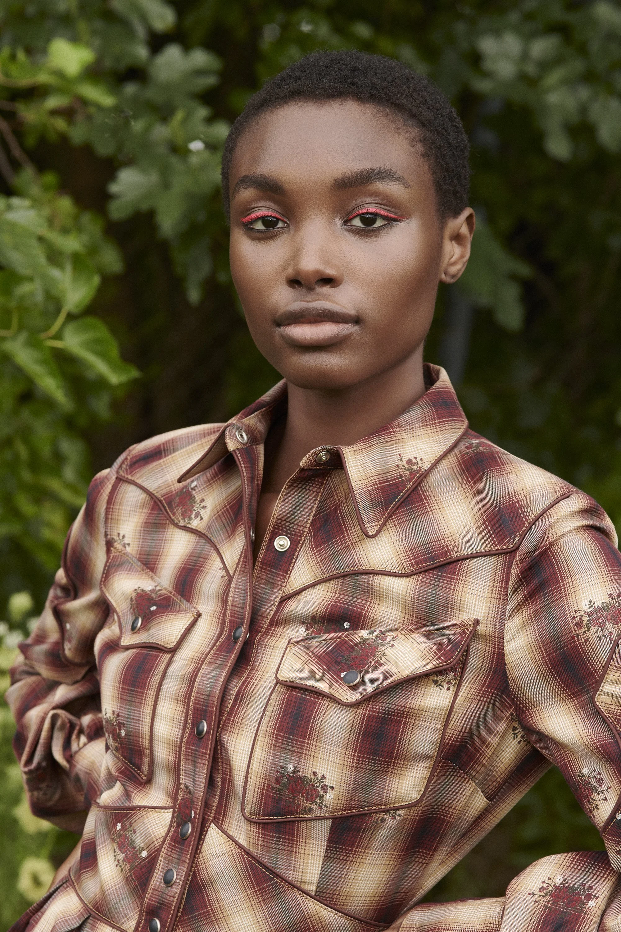 A young woman with short hair wearing a plaid shirt with floral patterns, standing outdoors with green foliage in the background.