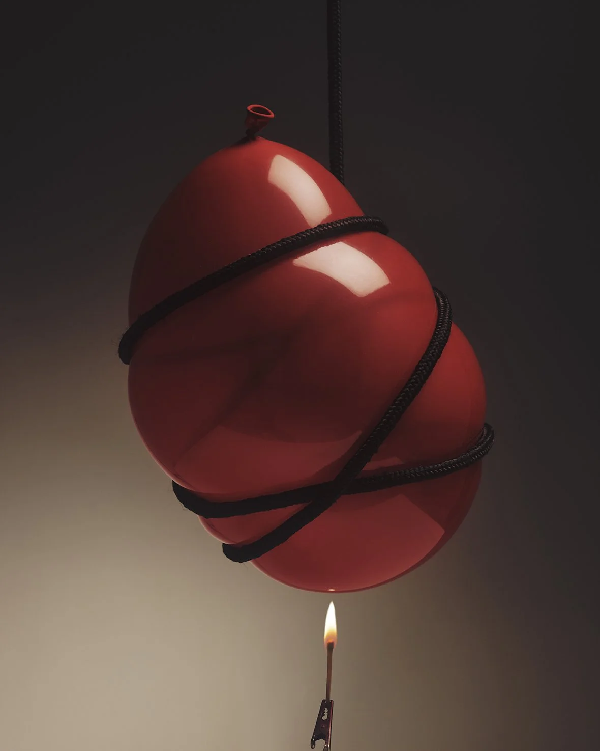 Two red balloons are tied together with black string, hanging upside down with a matchstick underneath lighting a small flame.