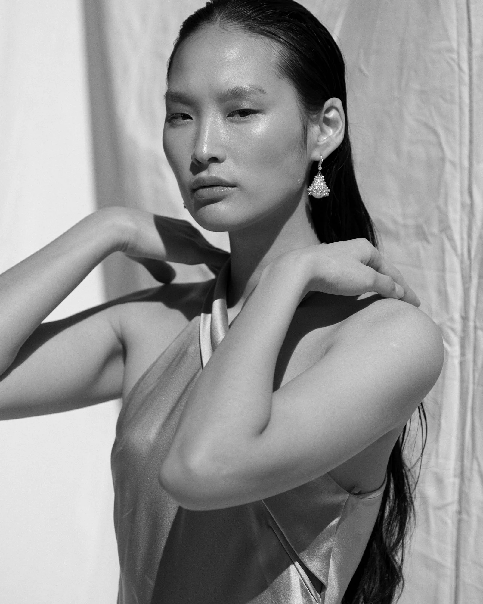 A black and white photograph of a woman with long, dark hair, wearing an earring and a satin top, posing with her hands on her shoulders against a plain backdrop.