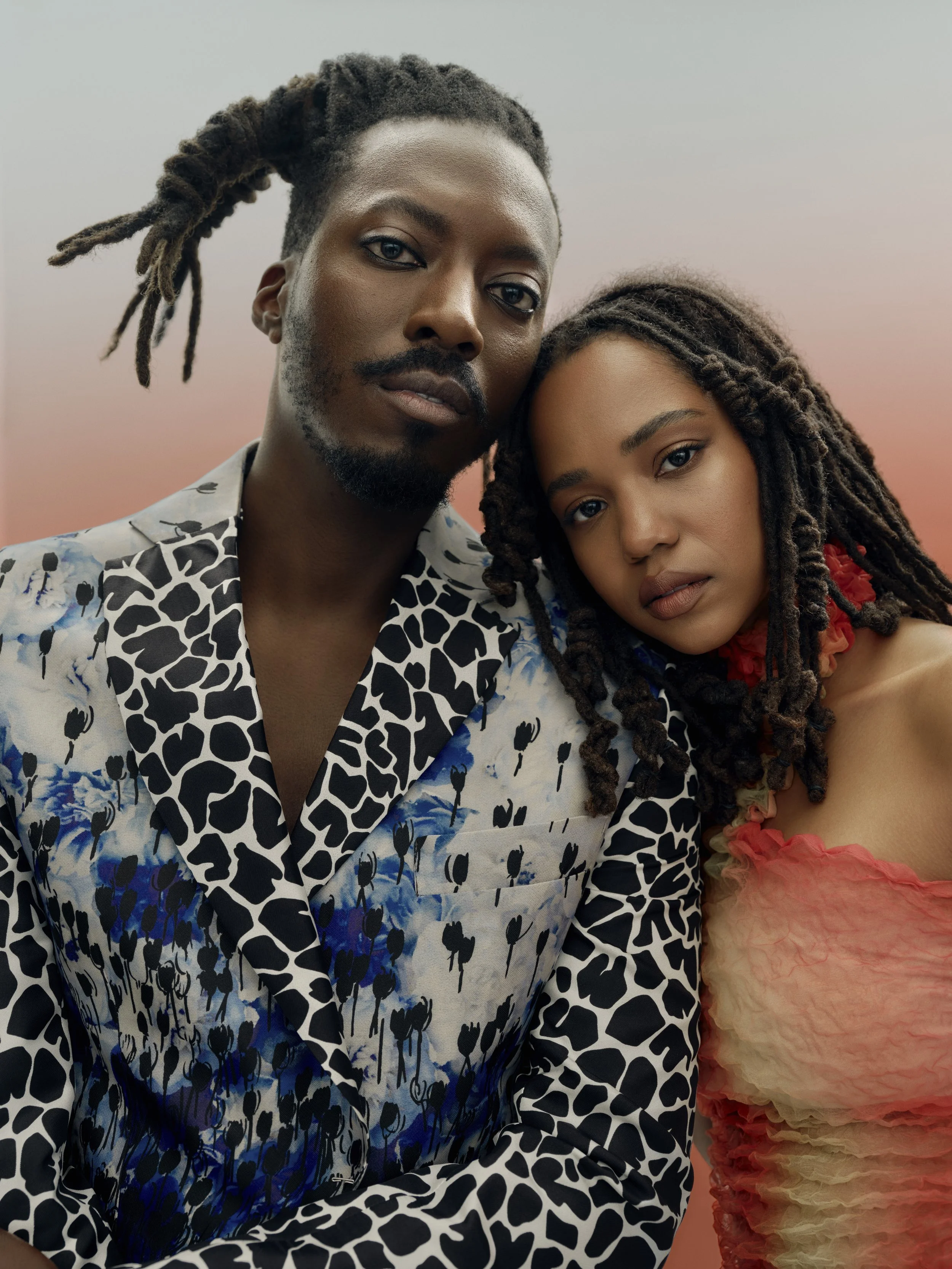 A close-up of a man and woman, both with dark skin and dreadlocks, posing together against a soft pink sky background. The man wears a patterned black and white shirt with blue floral accents, and the woman wears a strapless ruffled dress in shades o