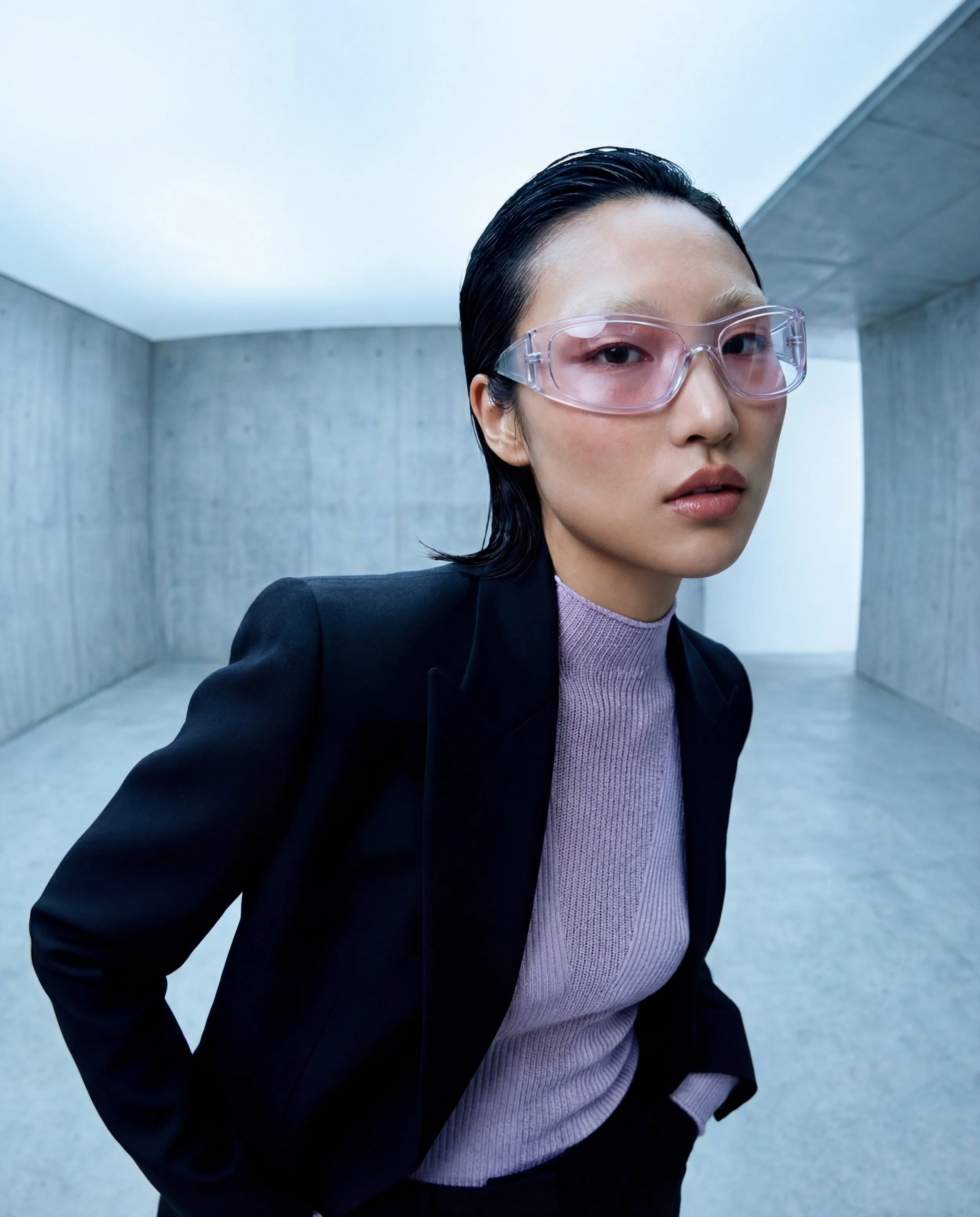 A woman in professional attire with short dark hair, wearing clear protective glasses, standing in a modern concrete indoor space.
