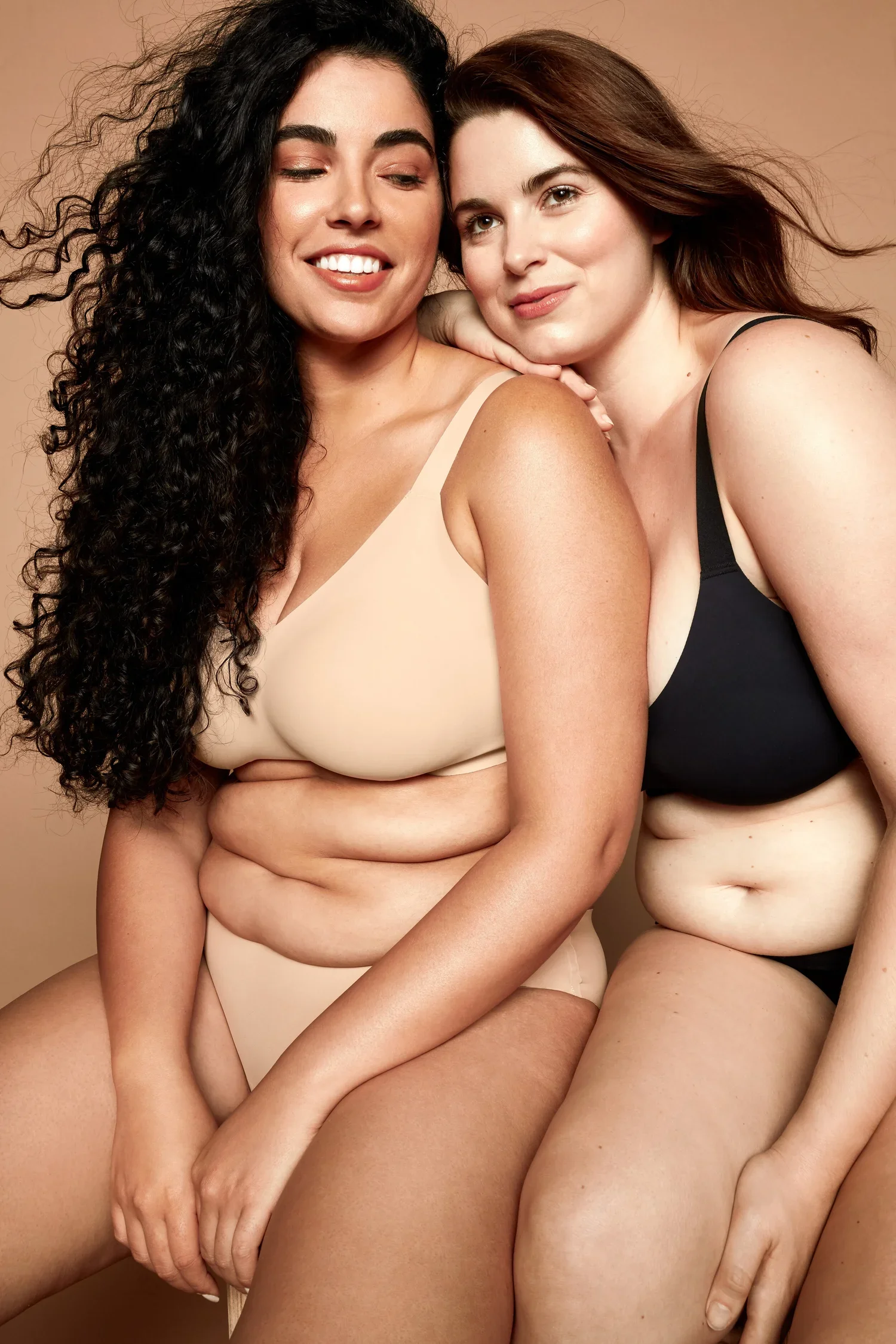 Two women sitting close together, smiling, with one resting her head on the other's shoulder. They are wearing nude-colored and black bras and underwear, against a beige background.