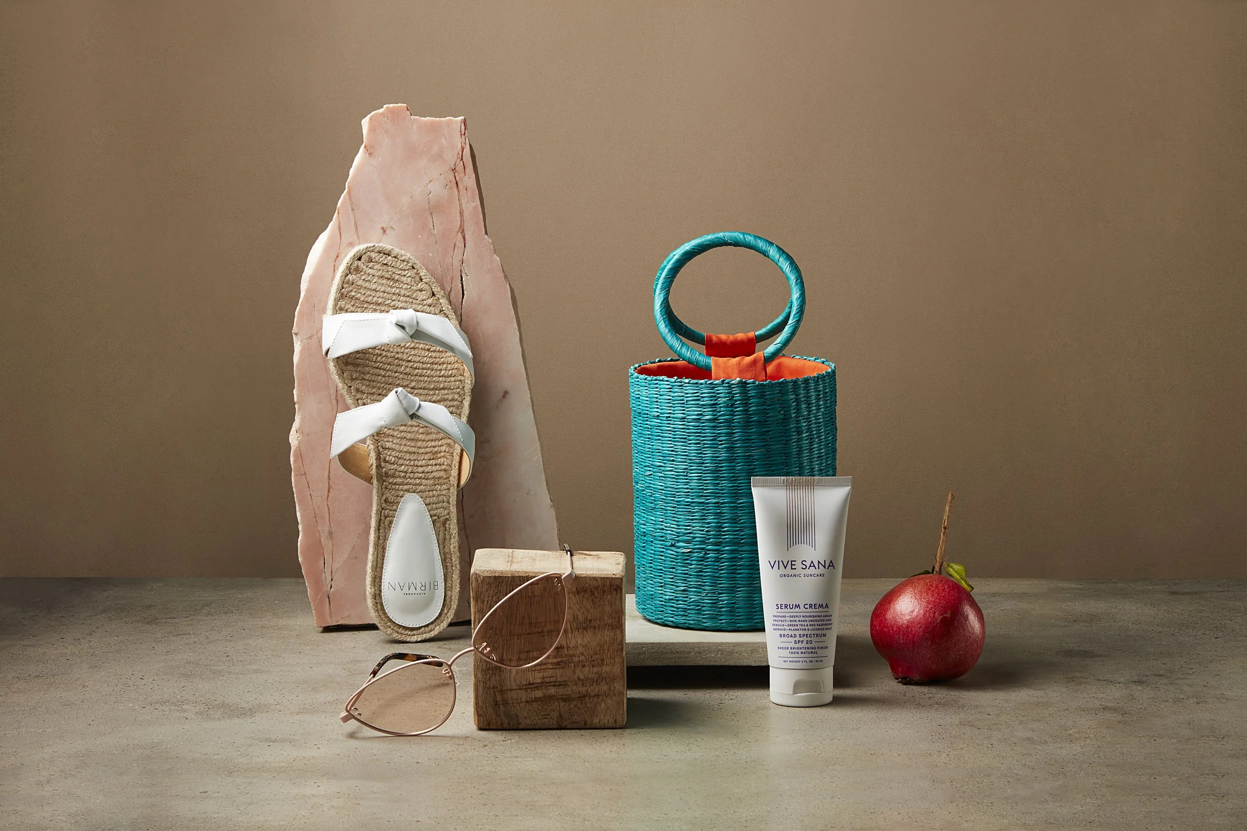 A flat lay of summer objects including a pink stone, a pair of beige sandals with white straps, a pair of sunglasses, a small wooden block, a blue woven basket with orange lining, a tube of Vive Sana skincare cream, a red apple, and a rock background