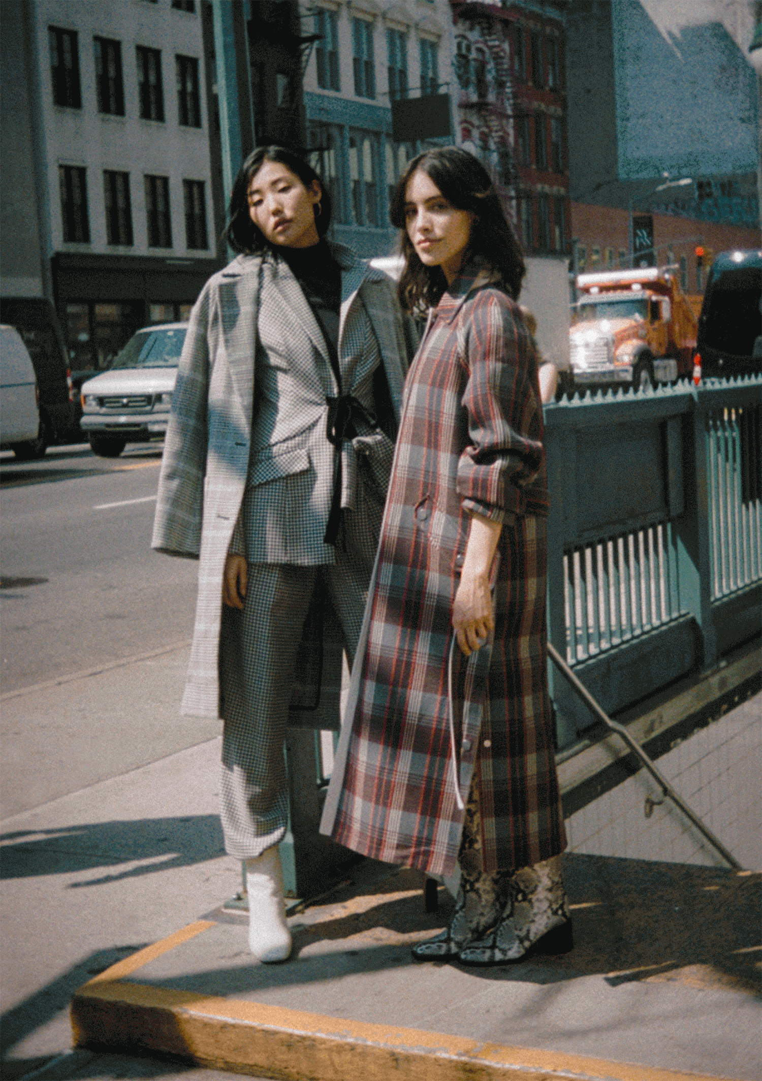 Two women standing on a city sidewalk in front of a railing with parked cars and buildings in the background, wearing stylish plaid and checkered coats.