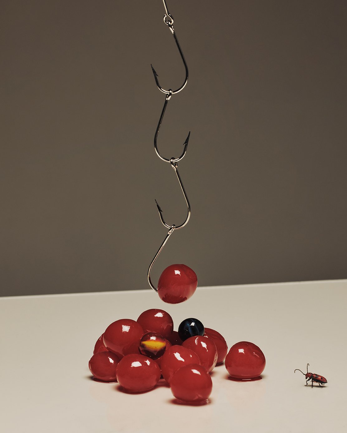 A fishing hook hanging above red and black jelly beans on a white surface, with a small toy insect to the right.