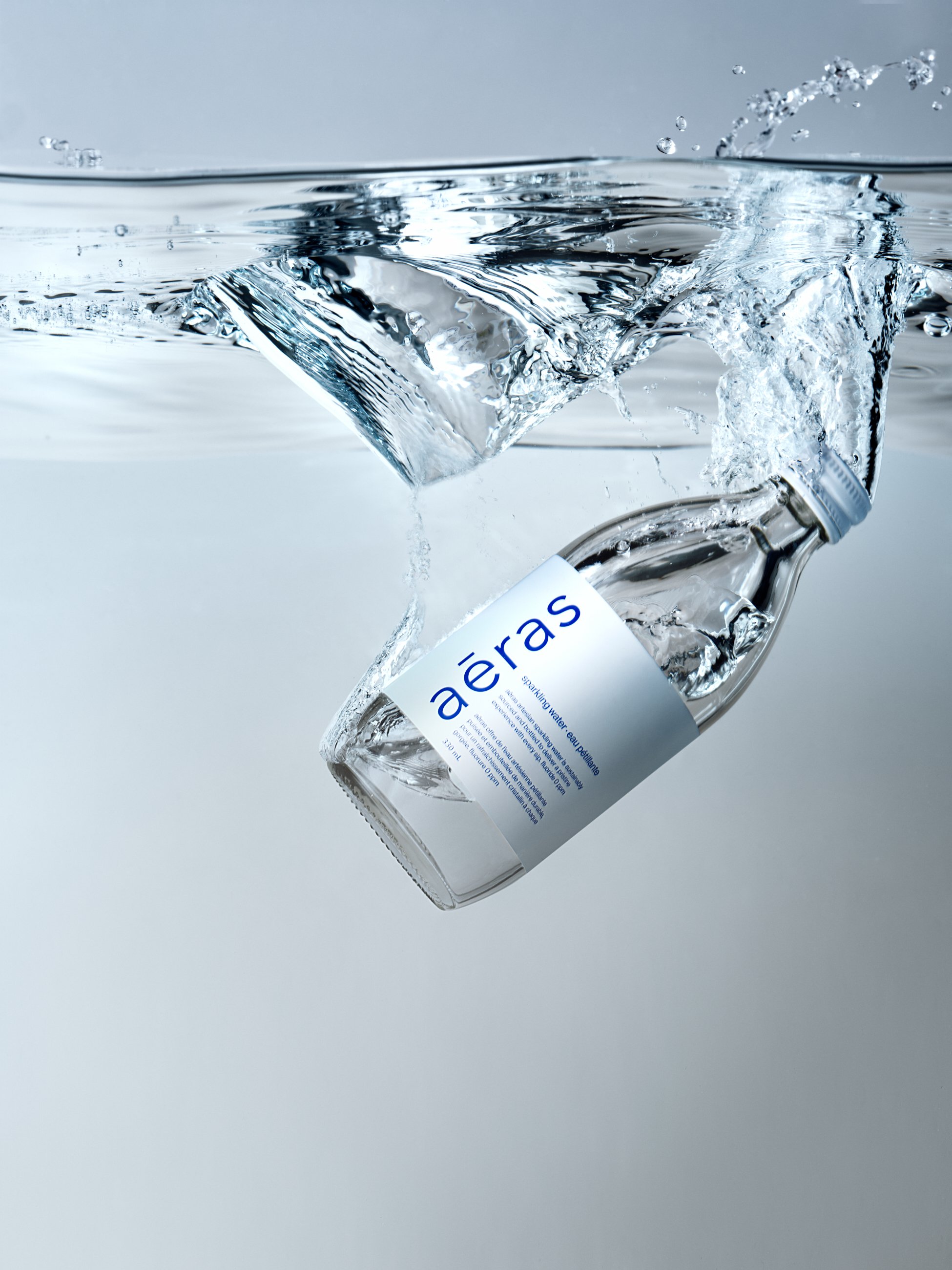 A clear glass bottle with a white label and blue text is falling into a body of water, creating splashes and bubbles. The water is transparent and the background is plain and light-colored.