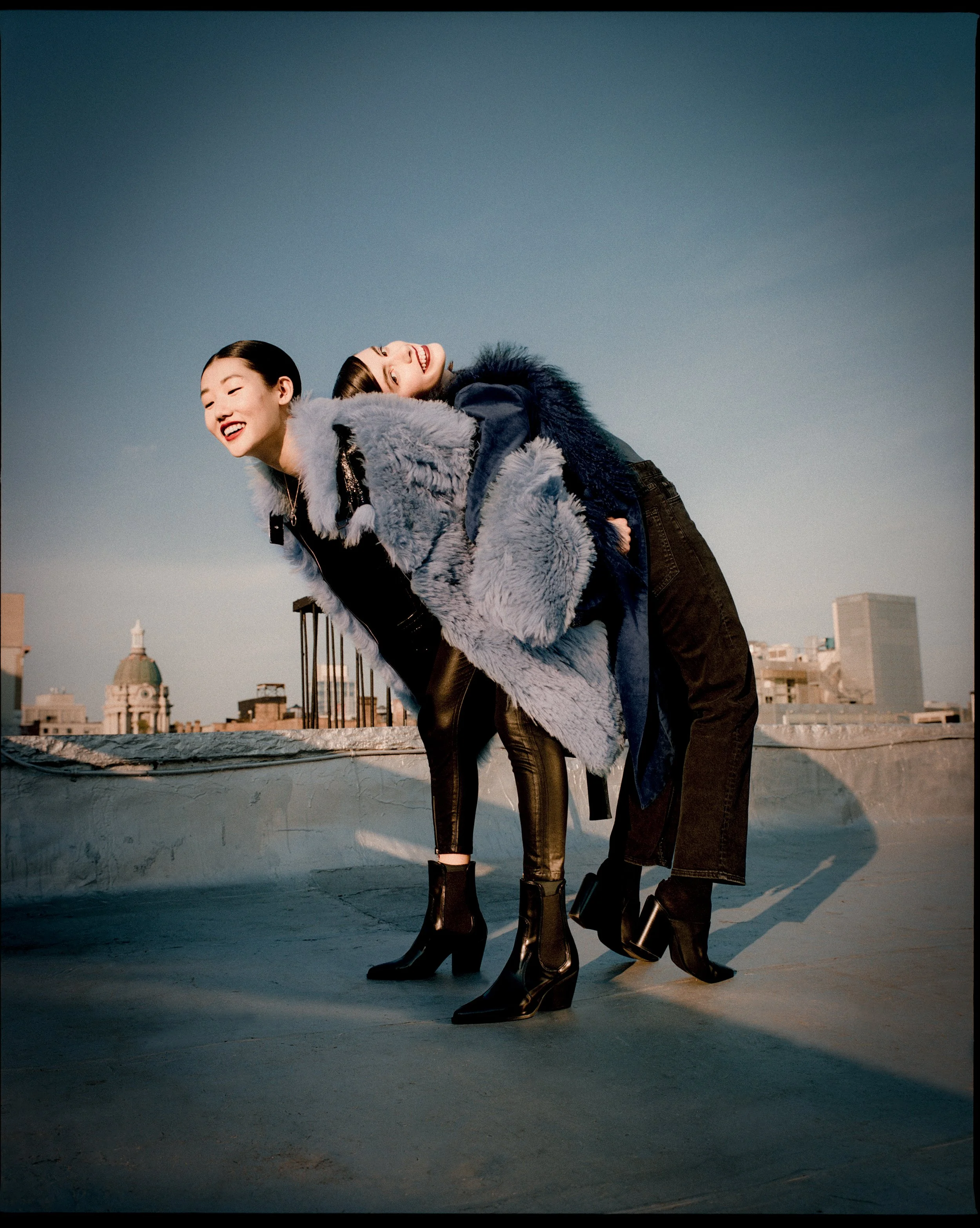 Two women with short dark hair smiling and leaning on each other on a rooftop, wearing fashionable winter clothing and high-heeled boots, with city buildings in the background under a clear sky.