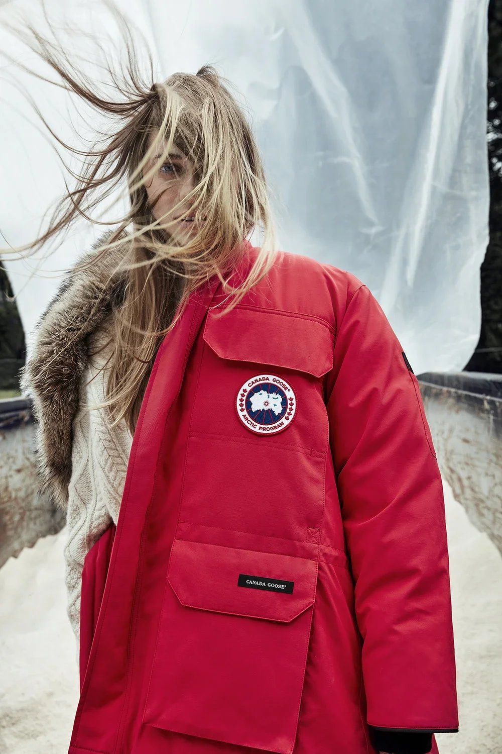 A woman with blonde hair blowing in the wind, smiling, wearing a red Canada Goose Arctic Program jacket.