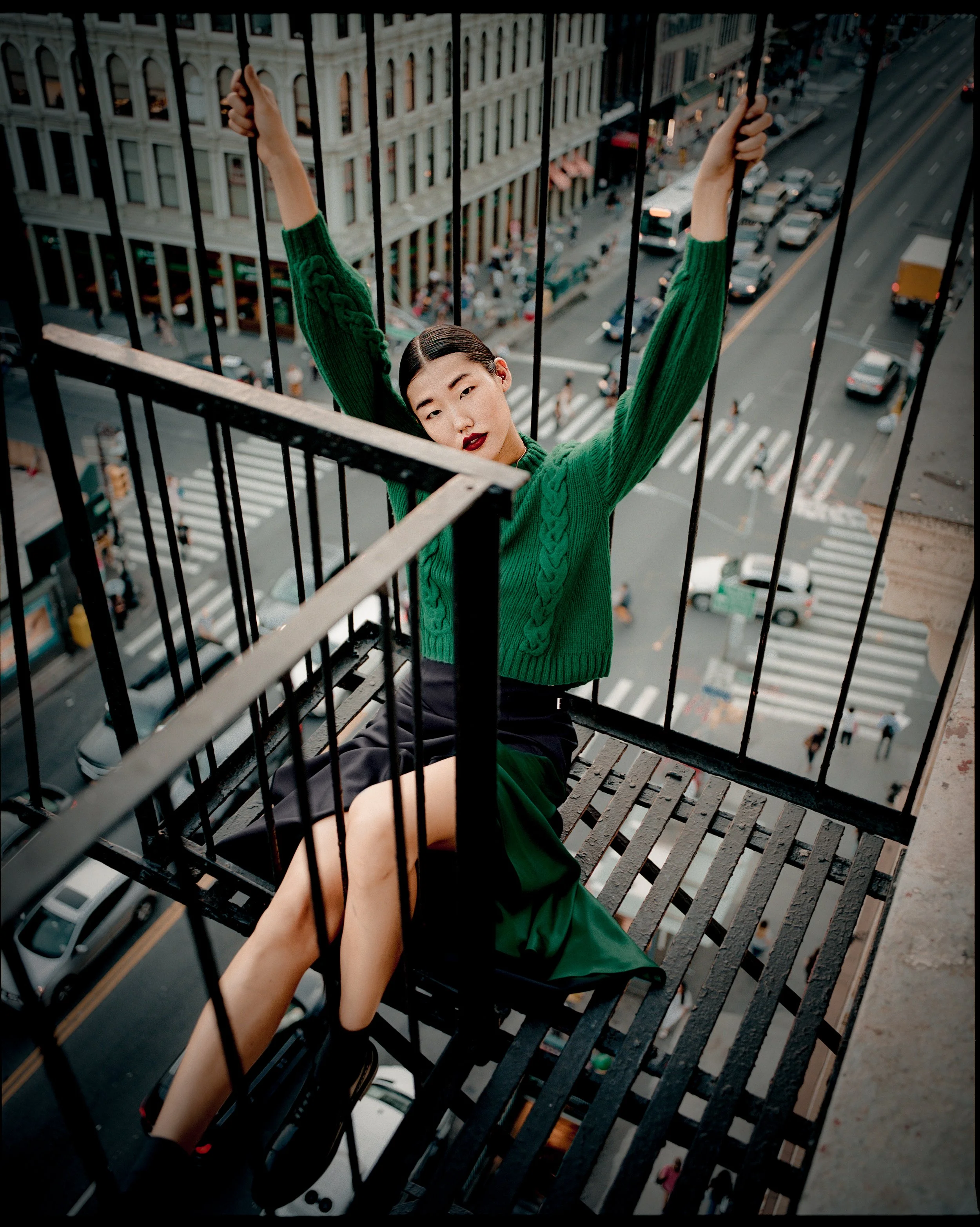A woman with dark hair and red lipstick, wearing a green sweater, sitting on a fire escape with her arms raised, overlooking a busy city street with cars and pedestrians.