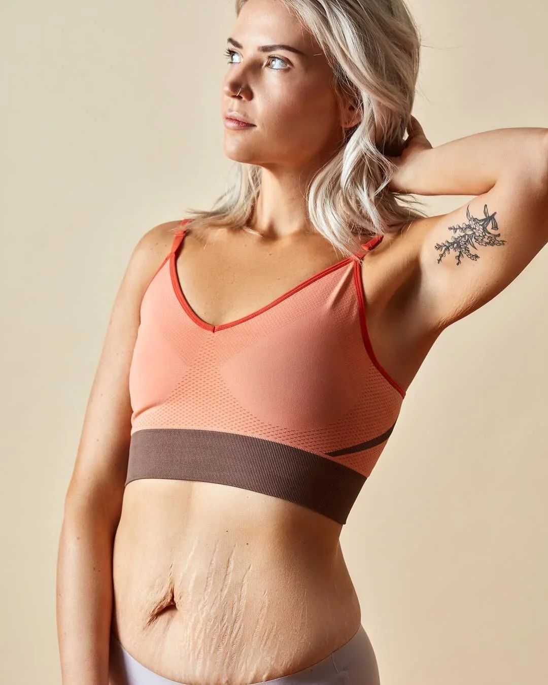 A woman with blonde hair and a tattoo on her upper arm wearing a peach-colored sports bra with red trim, standing against a beige background with her hand behind her head.