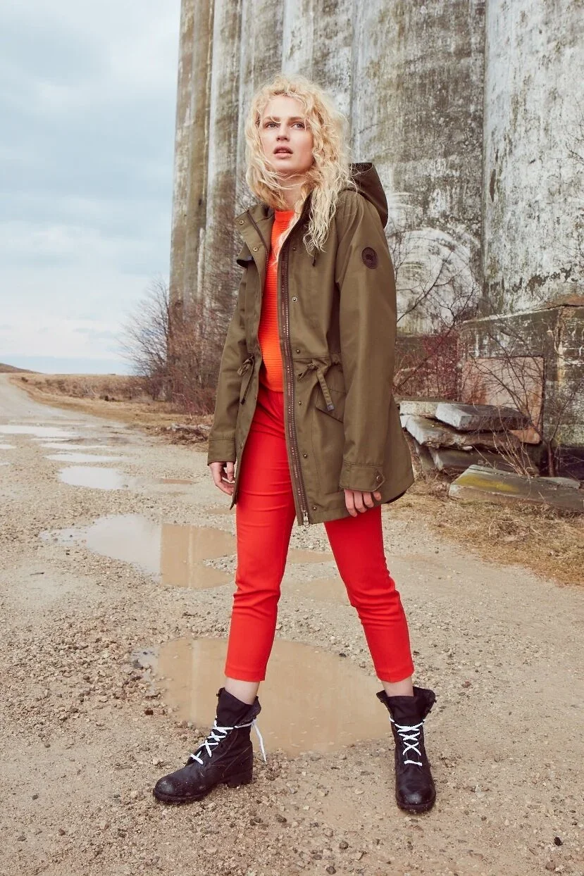 A woman with curly blonde hair standing outdoors on a dirt road with puddles, wearing a brown jacket, red pants, black boots, and an orange shirt, in front of a large weathered concrete silo.