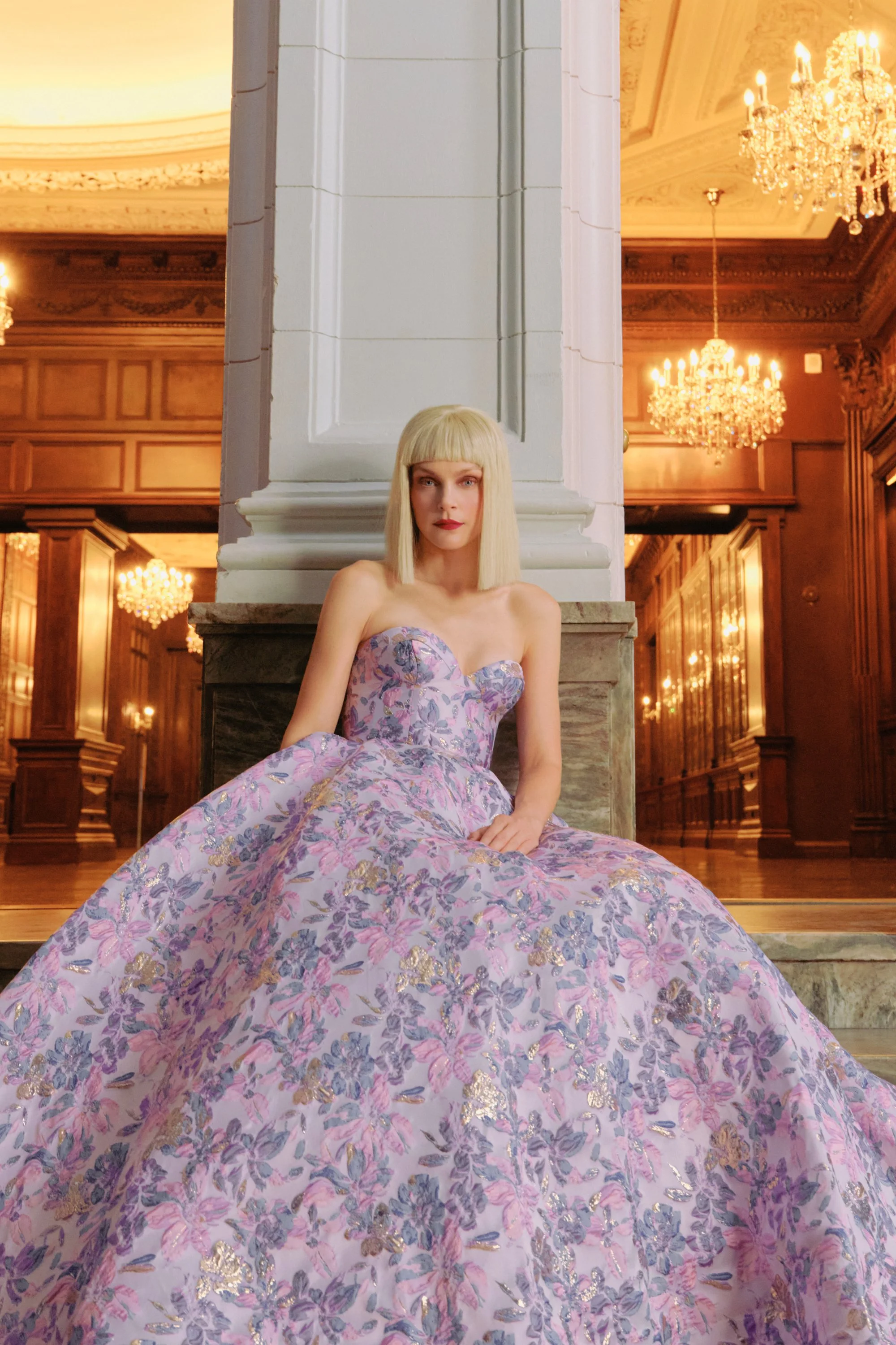 A woman with blond hair in a floral pink and purple ballgown sitting on the floor in an elegant, gold-lit room with chandeliers.
