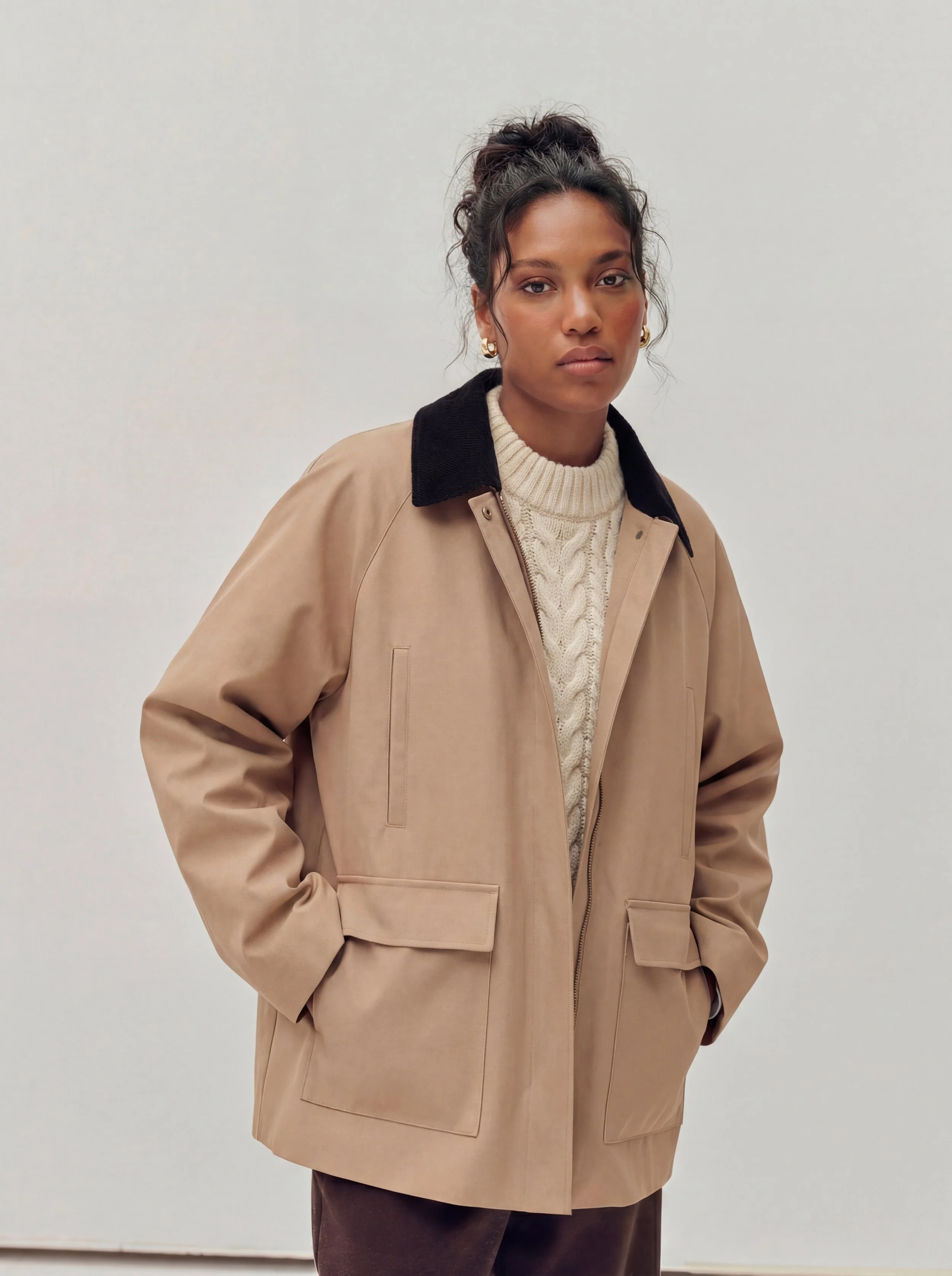 Woman wearing a beige jacket with black collar, cream cable-knit sweater, and gold earrings on plain white background.