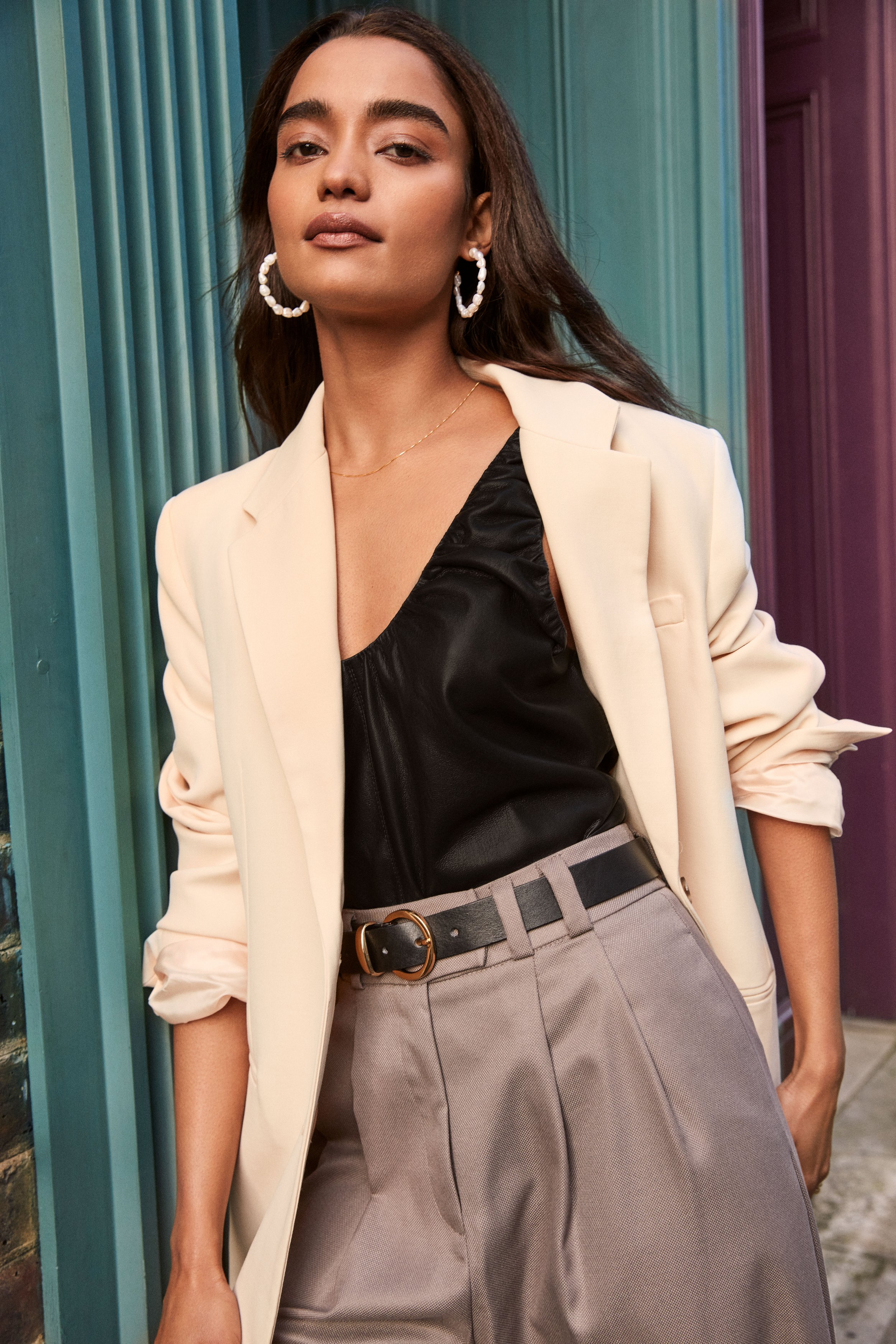 A woman with medium skin tone and long dark hair posing outdoors, wearing a cream blazer over a black satin top, gray high-waisted trousers with a black belt, pearl hoop earrings, a delicate gold necklace, and looking confidently at the camera.