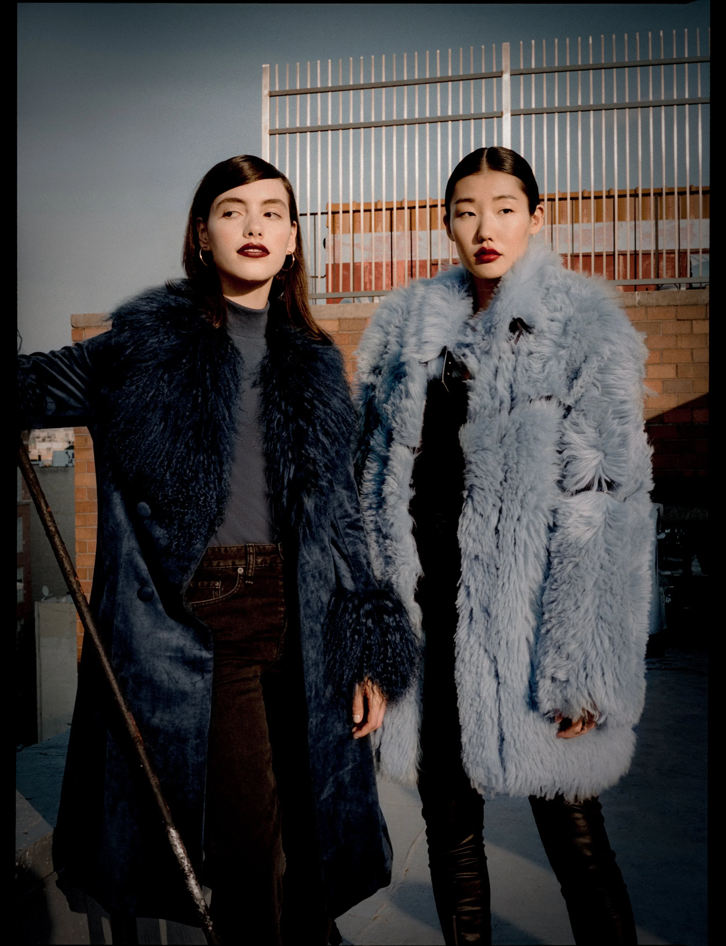 Two women wearing fur coats pose on a rooftop with a brick wall and a metal fence behind them.