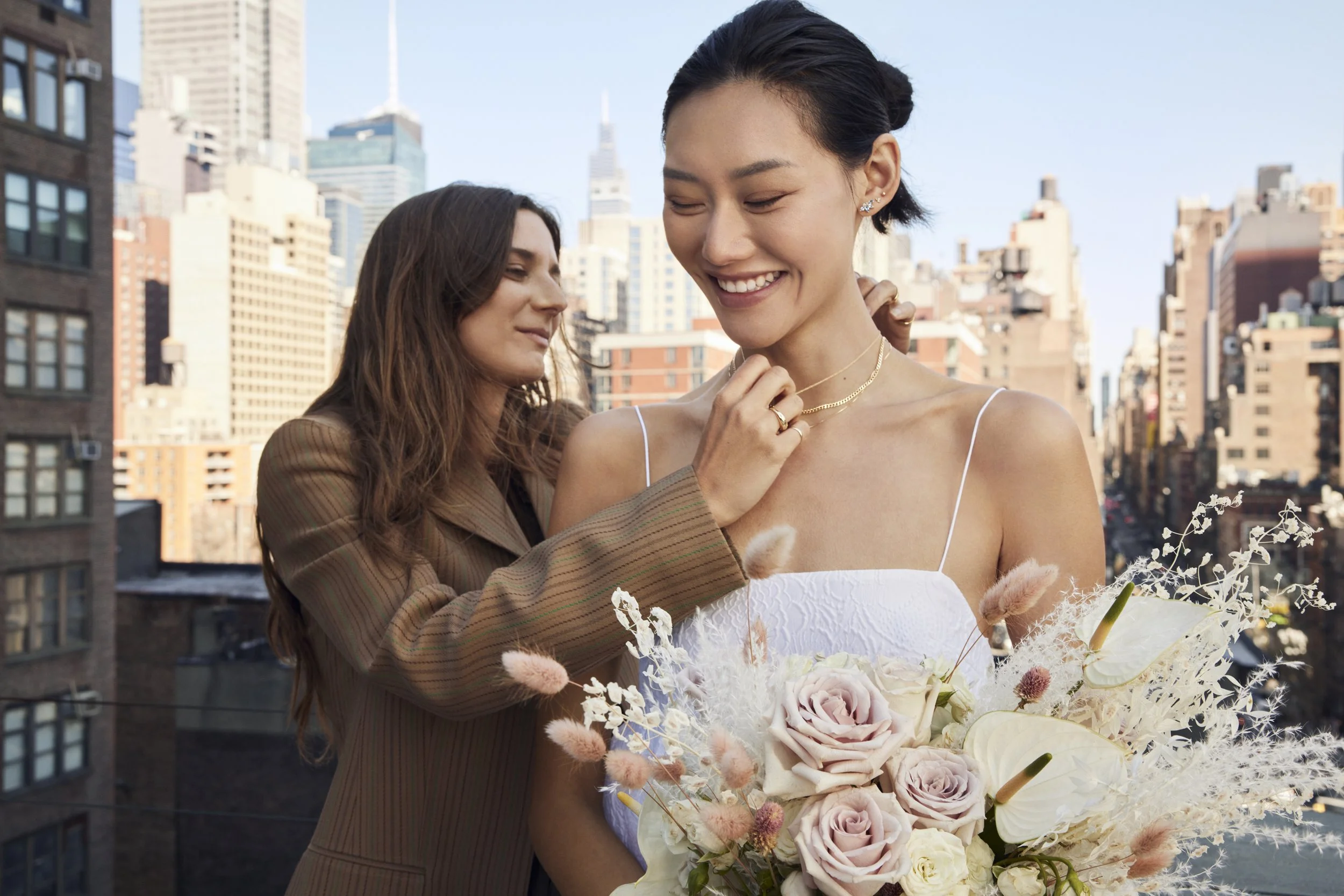 Two women, one helping the other with jewelry, standing outdoors in a city with tall skyscrapers, one woman holding a bouquet of pale pink roses and white flowers, smiling and wearing a white dress.