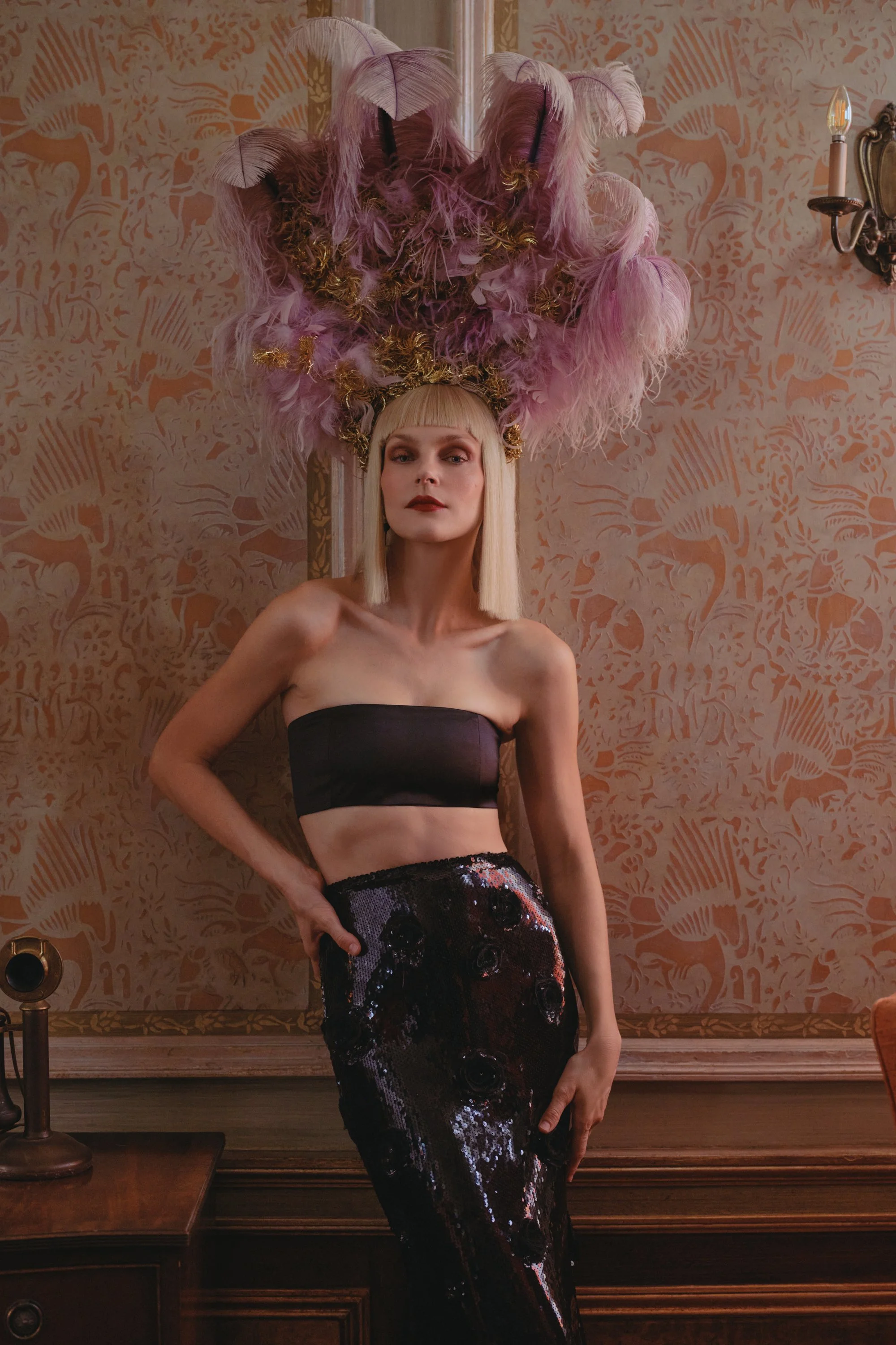 A woman with platinum blonde hair and bangs, wearing a strapless black top and a shiny black sequined skirt, standing against a patterned wallpaper in an elegant room with ornate decor, and a large pink feathered headpiece.