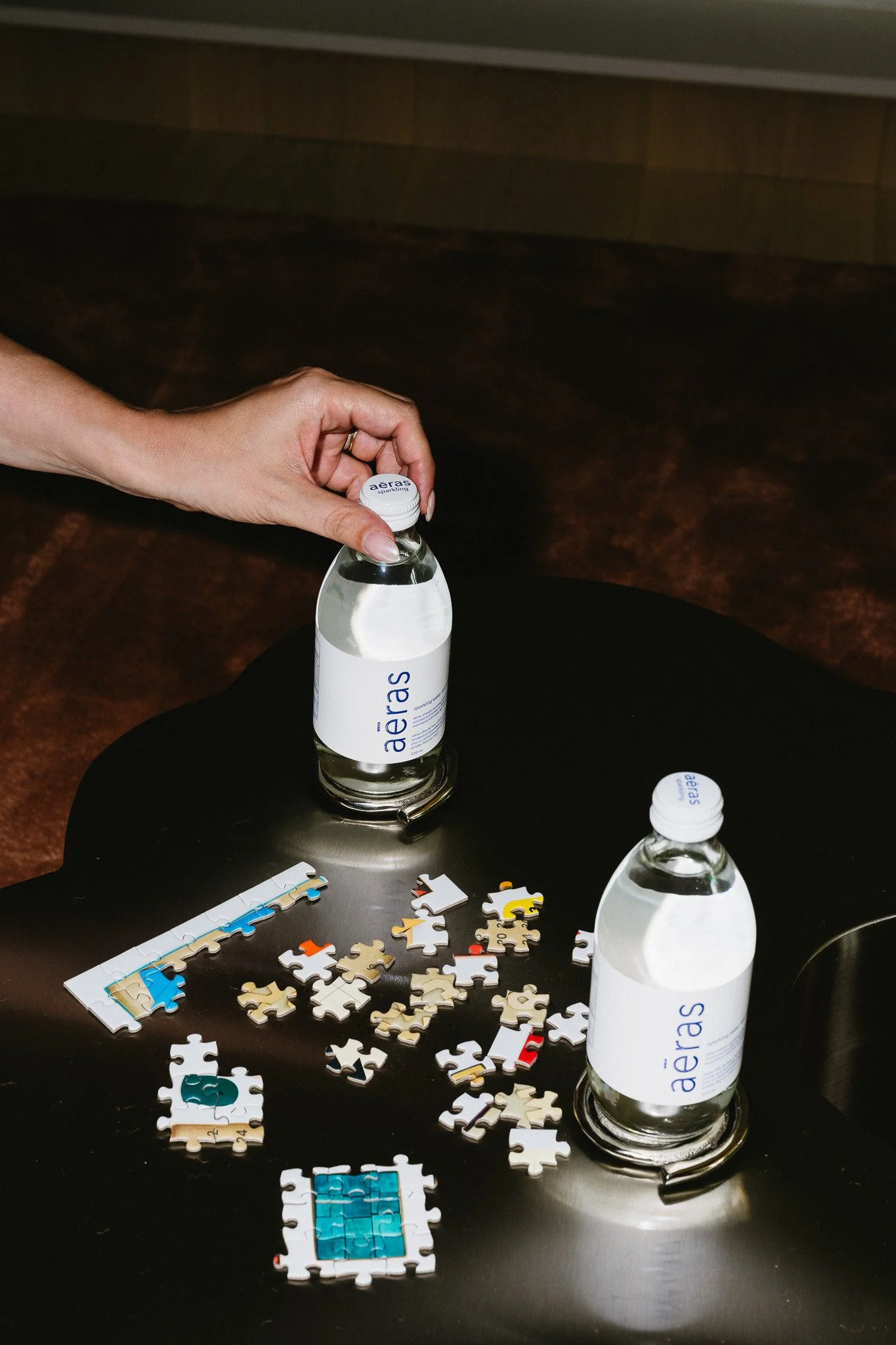 A hand reaching to open a bottle of water labeled 'aeras' on a dark surface with scattered missing puzzle pieces.