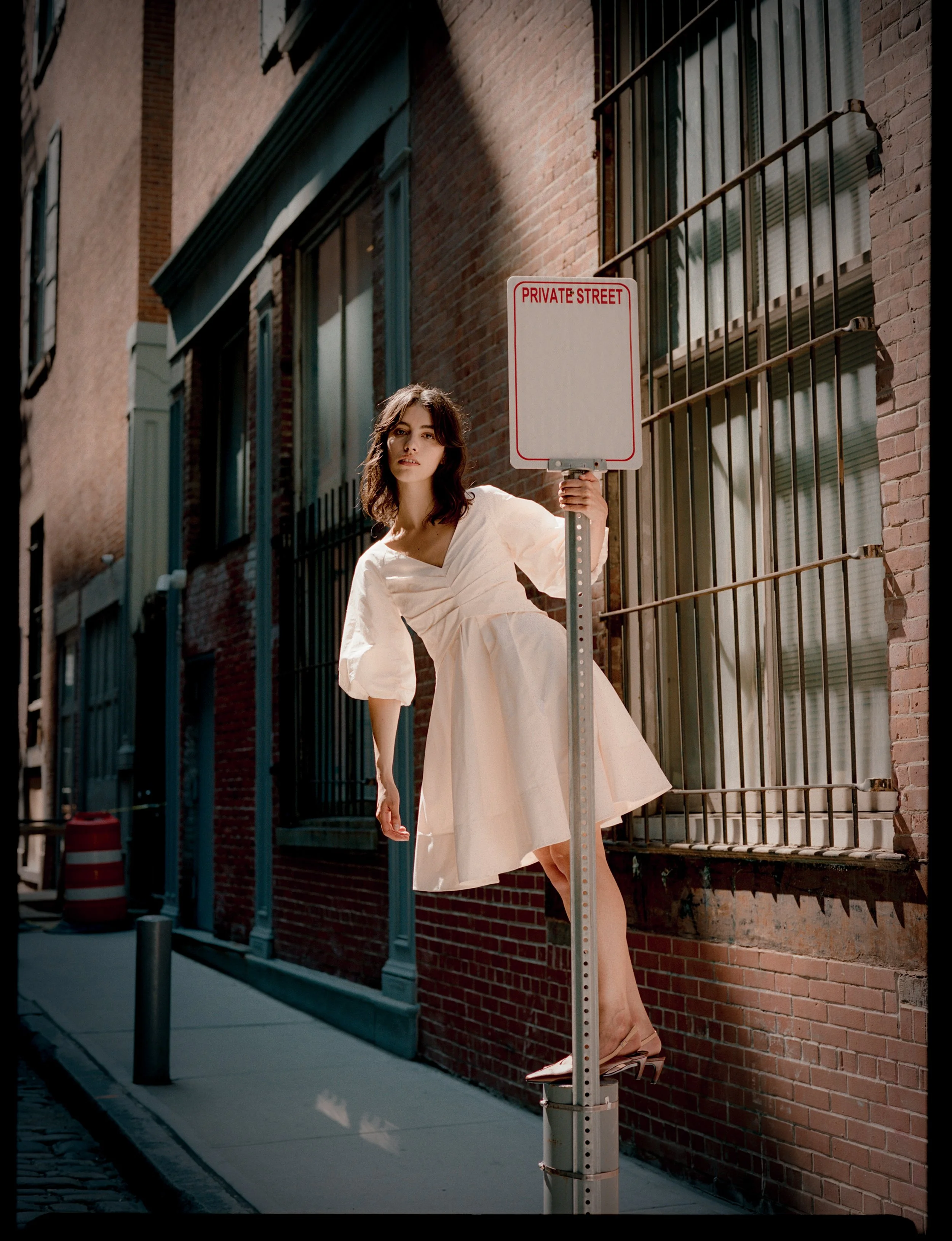 A woman standing on a fire hydrant, holding a 'Private Street' sign, in an alley with brick buildings.