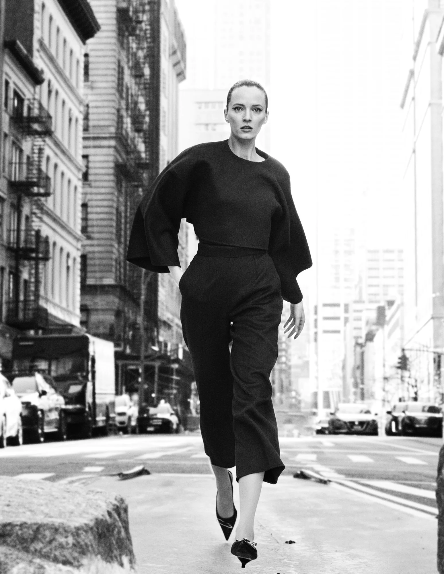 A woman walking on a city street in black clothing, with tall buildings and parked cars in the background.