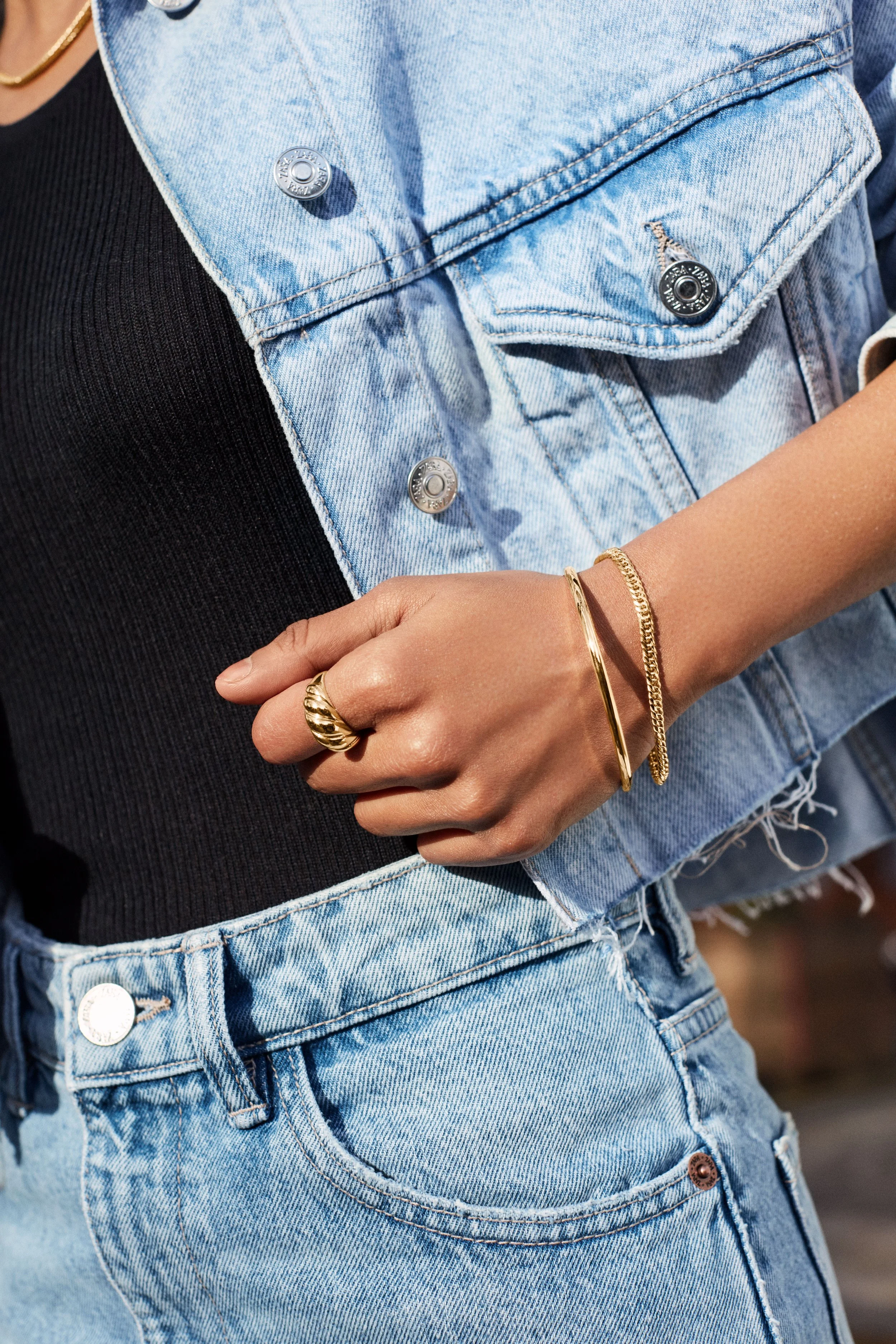 Close-up of a person wearing a light denim jacket over a black top, with gold jewelry including rings and bracelets, and light-wash jeans.