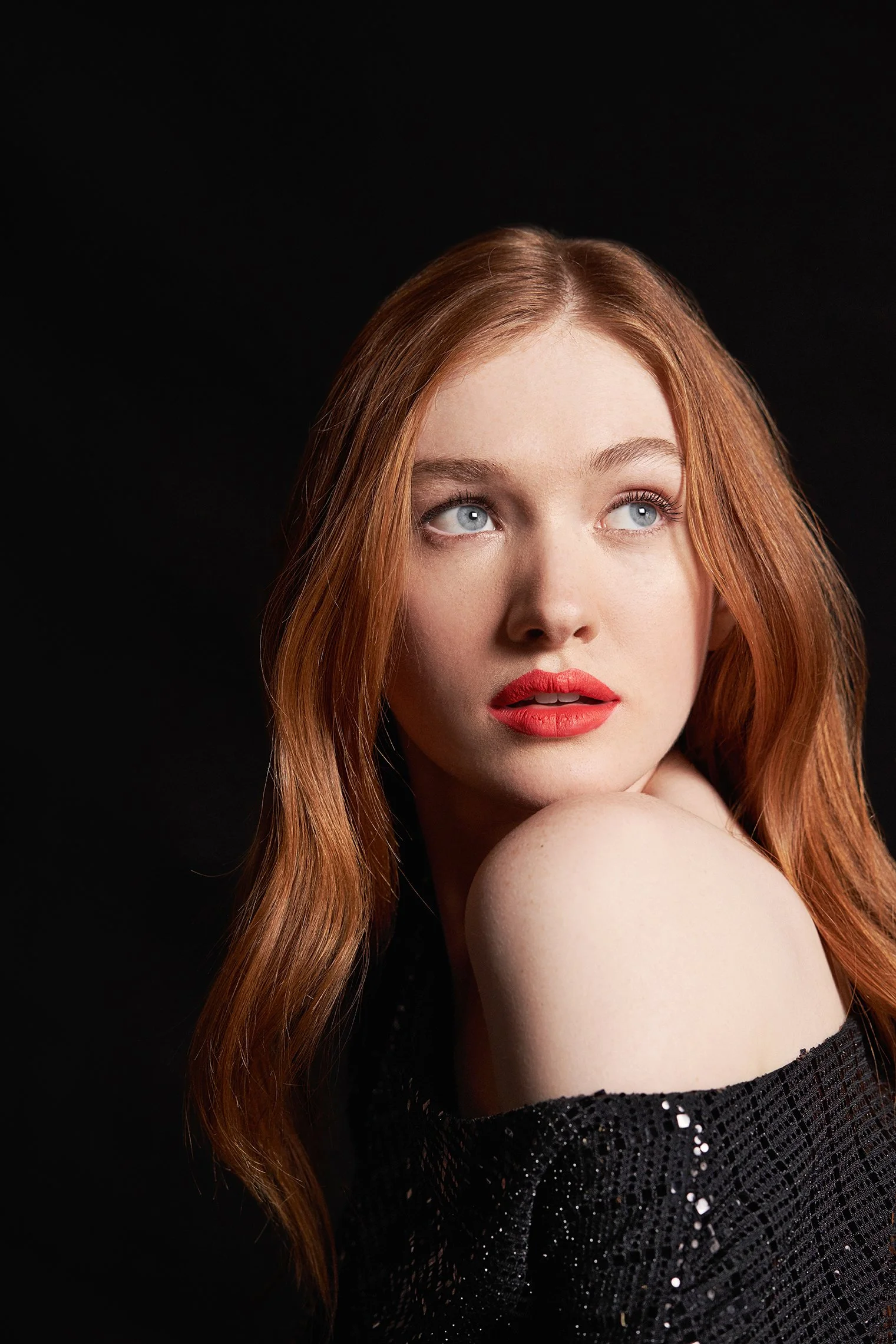 A woman with red hair and blue eyes, wearing red lipstick, looking slightly upward and to the side, against a black background.