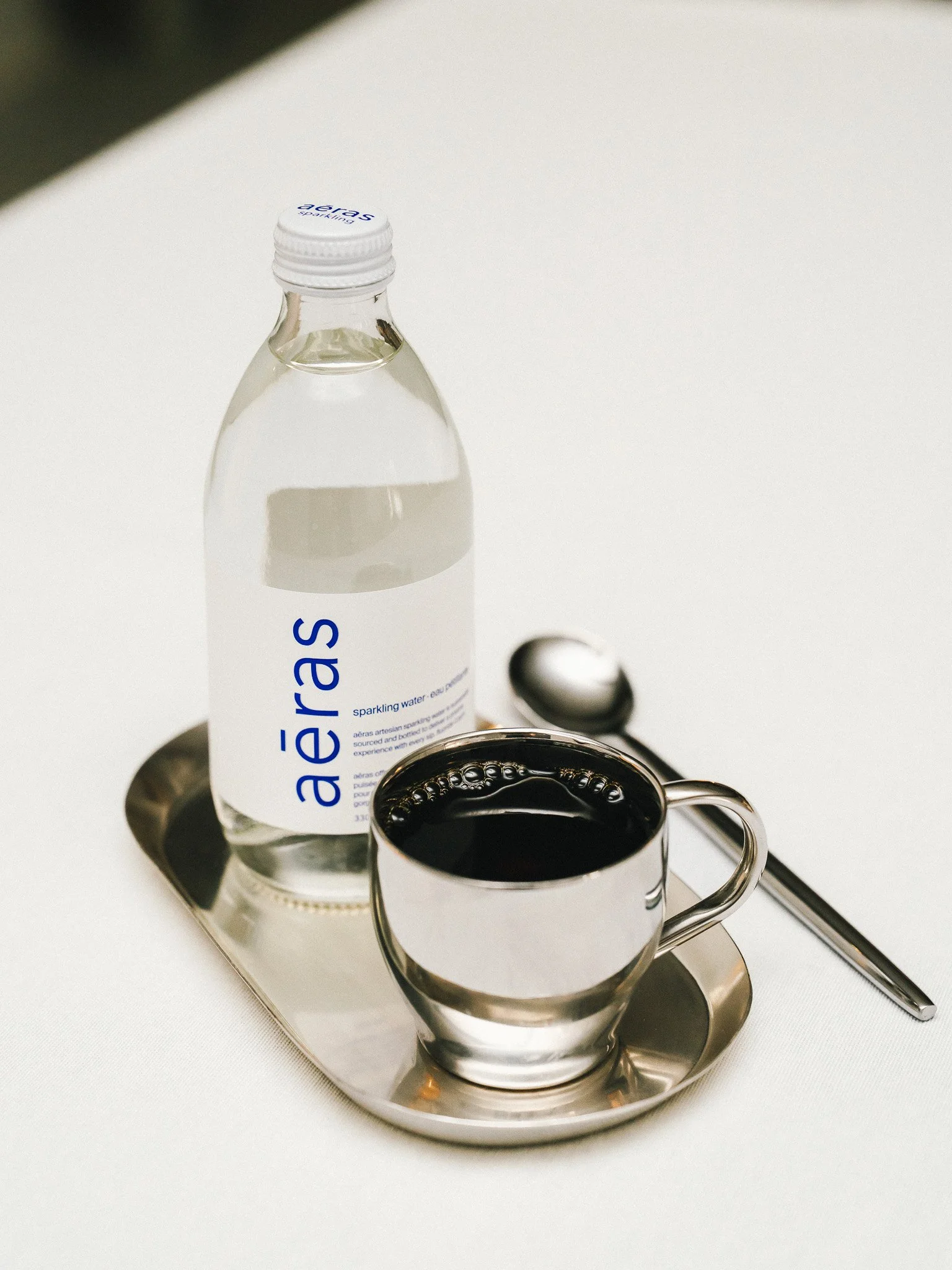 A bottle of sparkling water, a cup of black coffee, and a spoon on a silver tray on a white table.