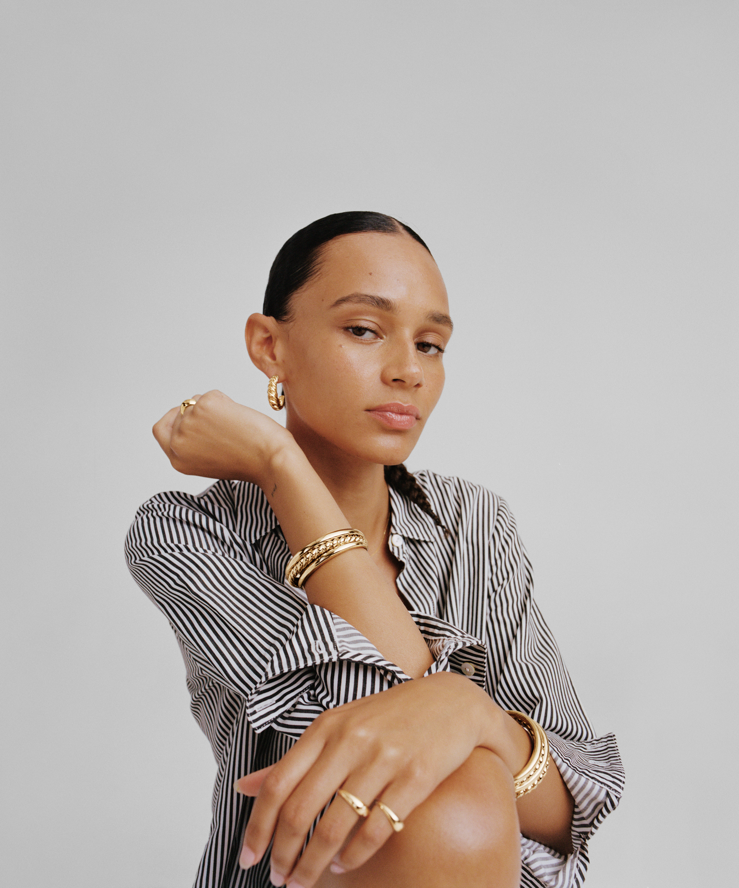 A woman with dark hair pulled back, wearing gold jewelry including earrings, bangles, and rings, posed with arms crossed, dressed in a black and white striped shirt, against a plain light gray background.