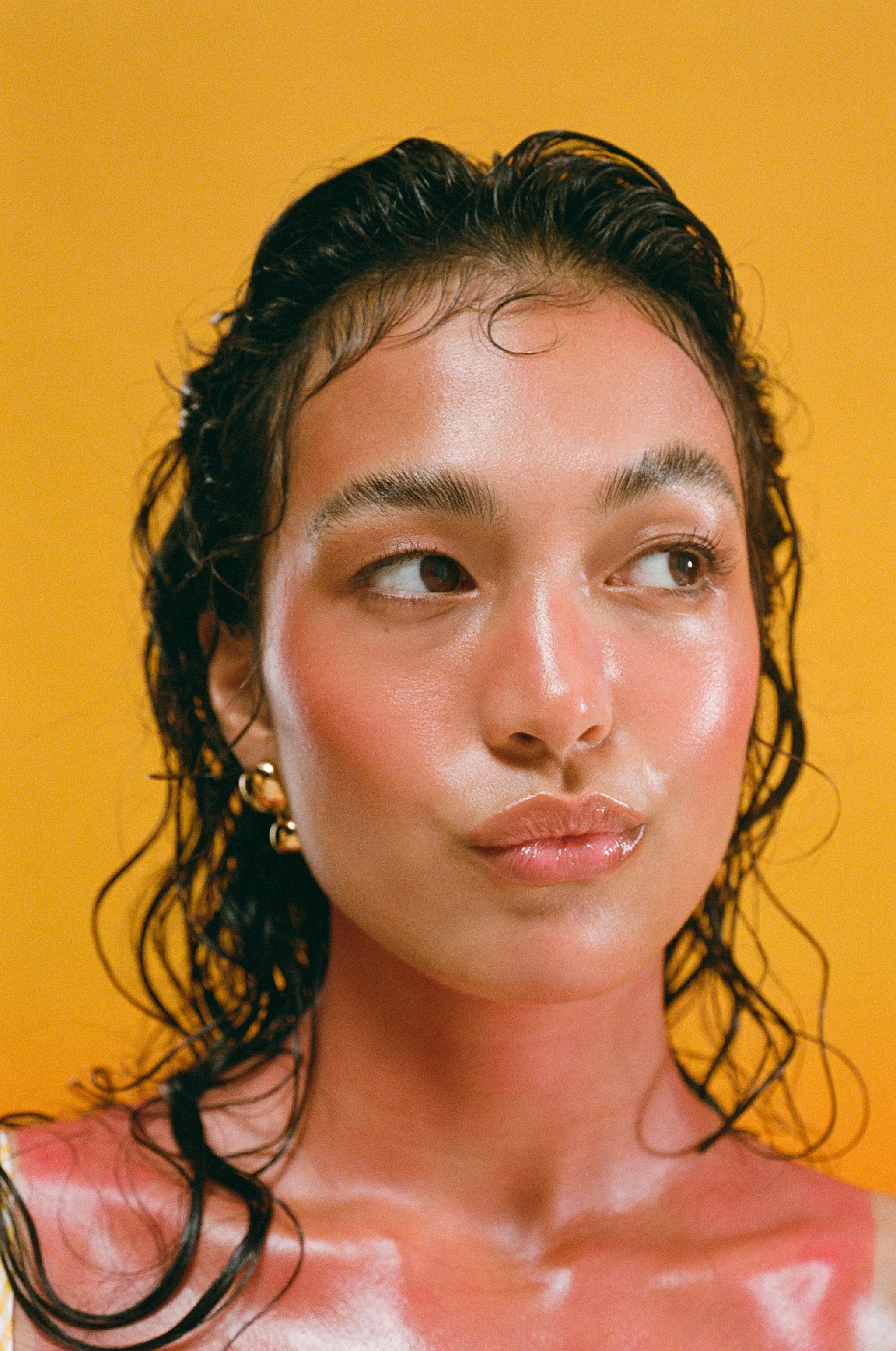 Close-up of a young woman with wet, curly hair against a yellow background, looking to the side with a calm expression.