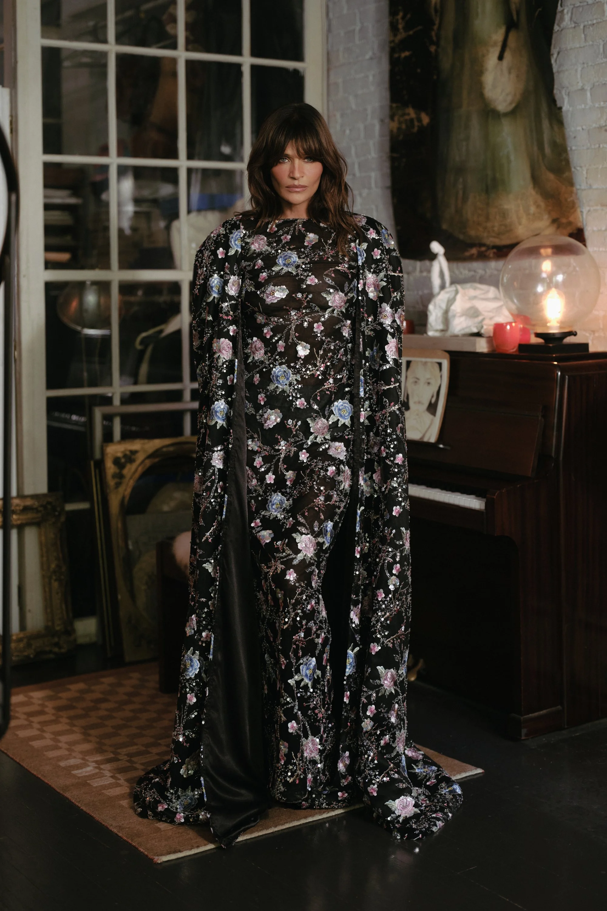 Woman in a long, floral, black velvet dress standing in a room with vintage decor and a piano.